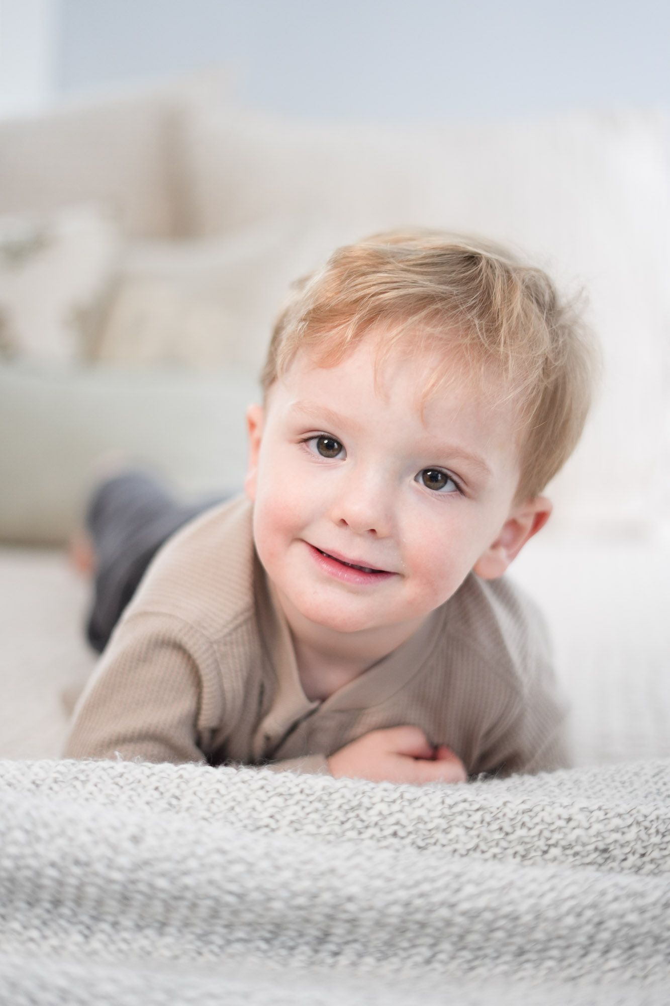 Happy toddler boy with light hair smiling while lying on a soft gray blanket.