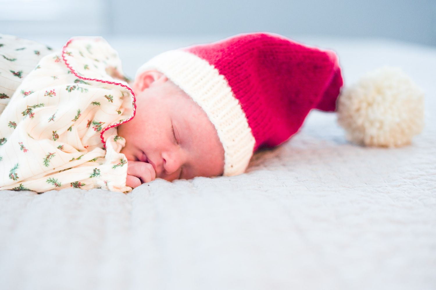 Peaceful newborn baby sleeping in Christmas onesie with red knit Santa hat on white surface.