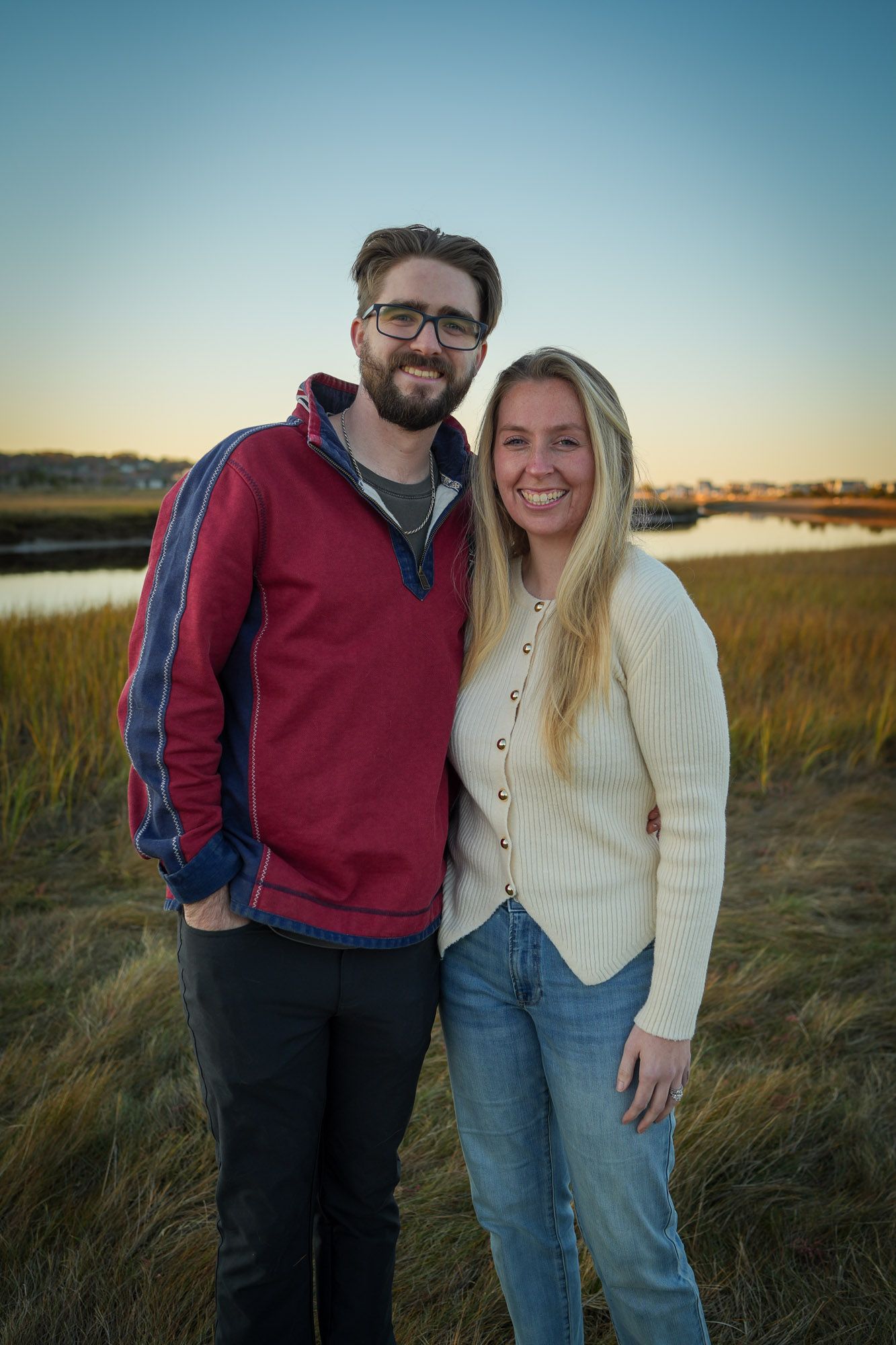 Smiling couple posing together by a marsh at golden hour, wearing casual sweaters and jeans.