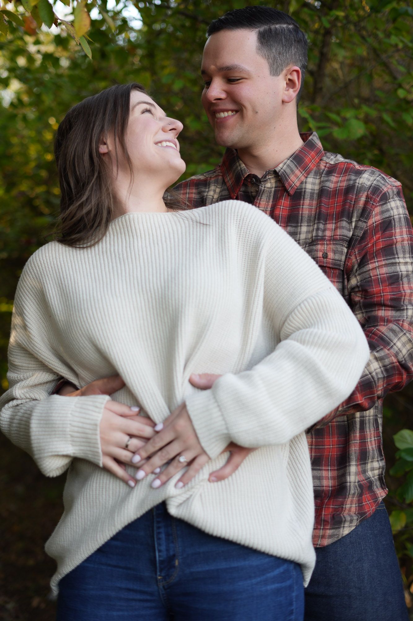 Smiling couple embracing outdoors surrounded by colorful autumn foliage, woman wearing engagement ring.