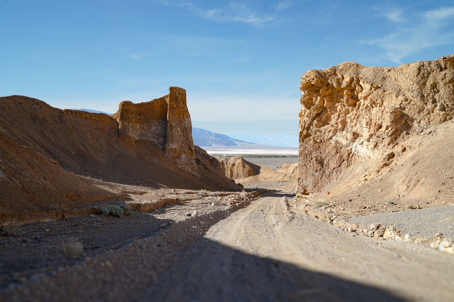 Narrow desert canyon with towering sandstone cliffs flanking a dusty dirt road under a clear blue sky.