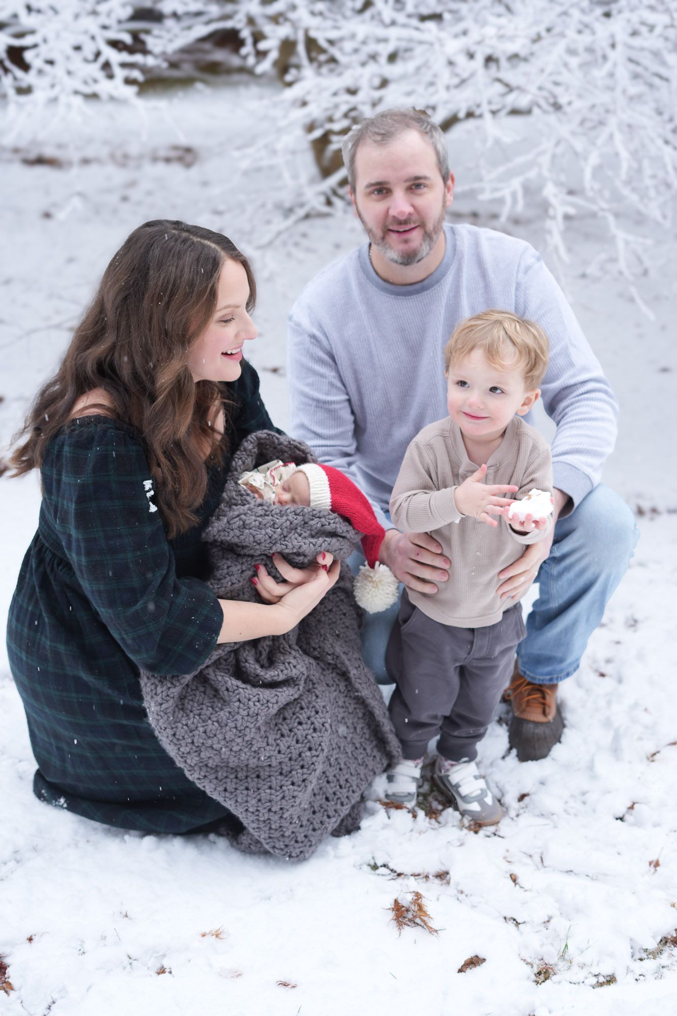 A family of four enjoys a snowy winter day outdoors, with parents smiling as their toddler son holds a snowball near the newborn.