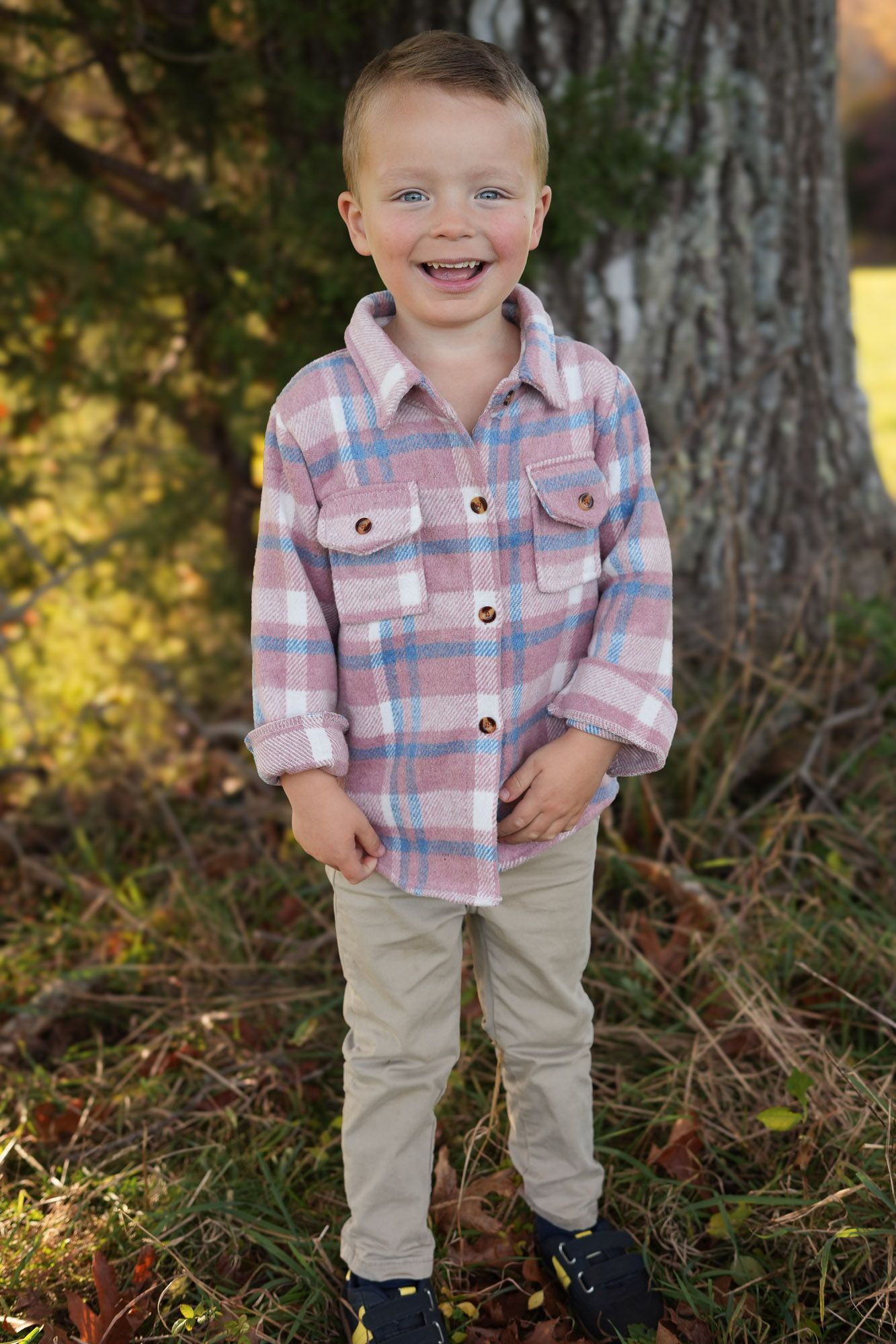 Cheerful young boy in a plaid flannel shirt smiling in front of a tree in autumn.