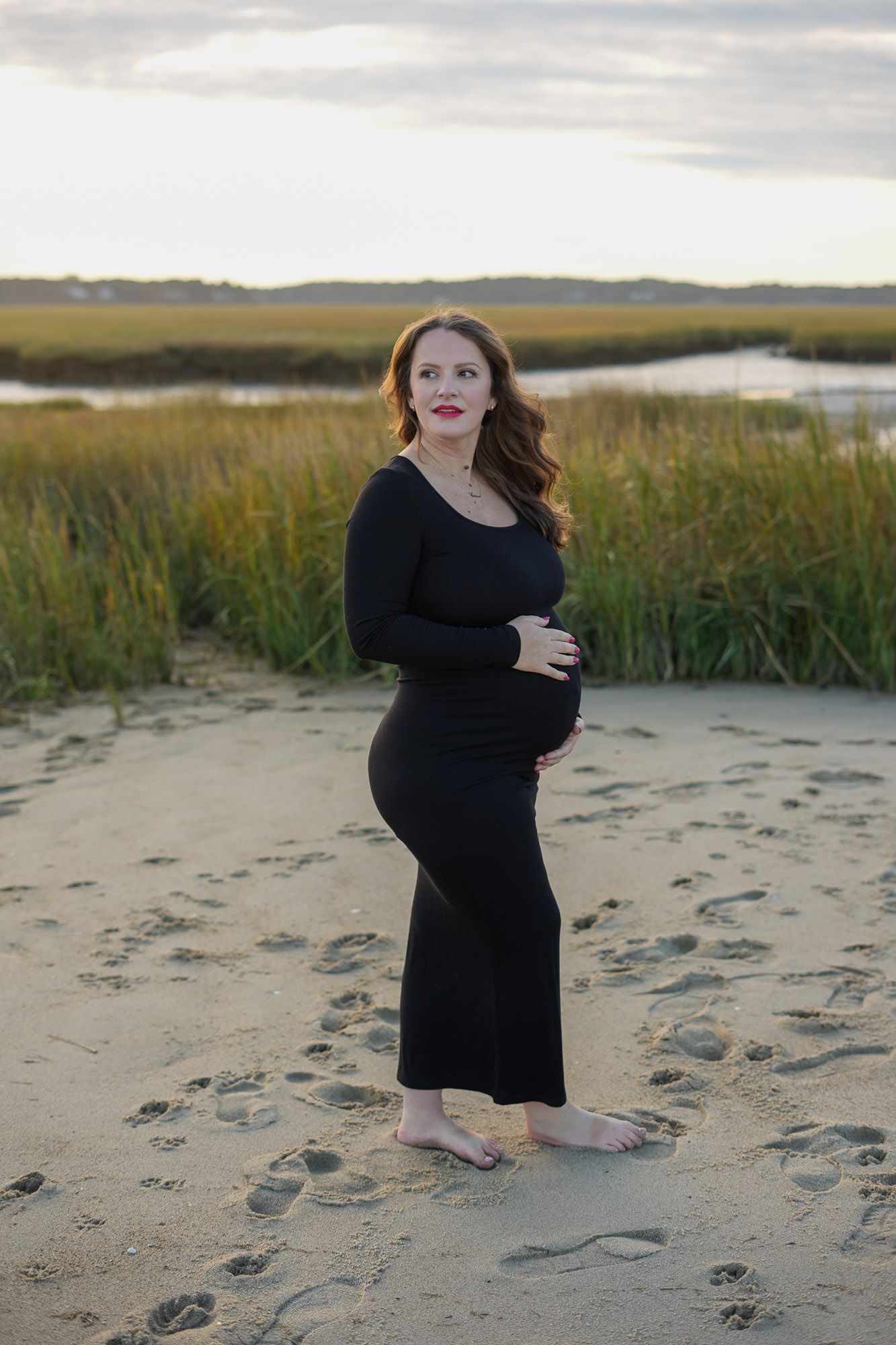 Pregnant woman in black maxi dress holding belly on sandy beach near marsh grass.