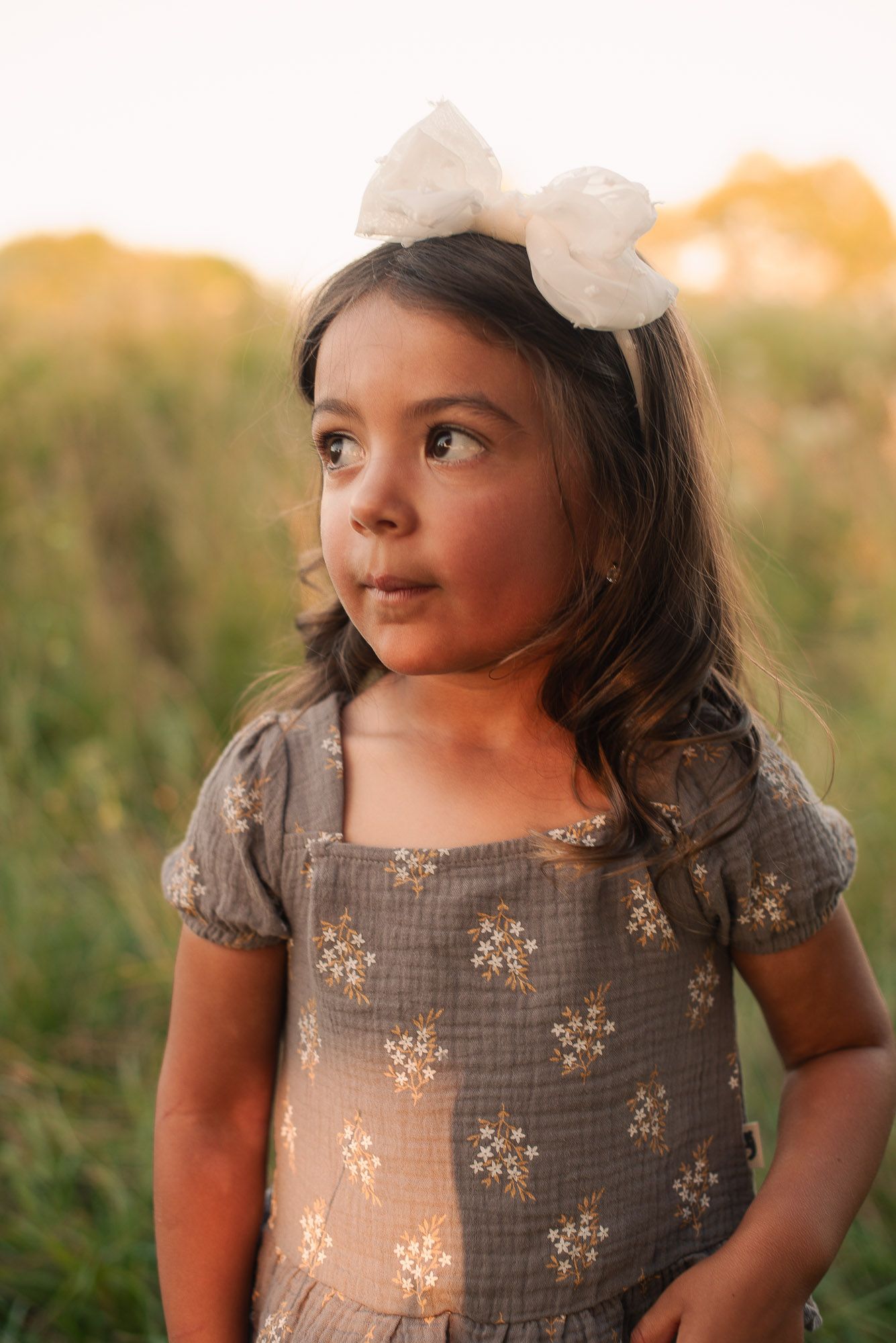 An adorable little girl in a floral dress and white bow headband gazes upward in a sunny field.