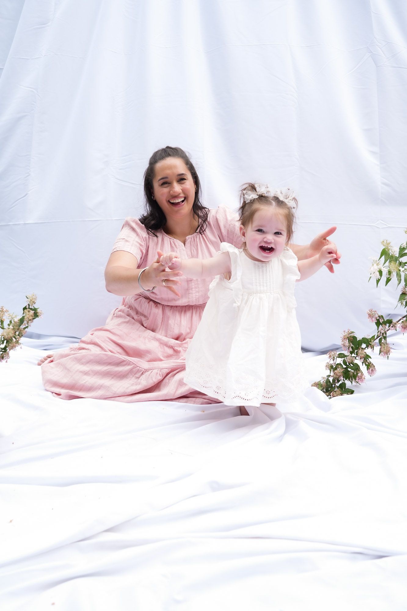 A laughing mother in pink reaches for her excited toddler daughter in a white dress during an outdoor photo session.