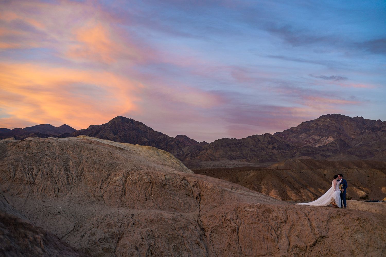 Bride and groom sharing a romantic kiss on rocky desert terrain at colorful sunset with mountains.