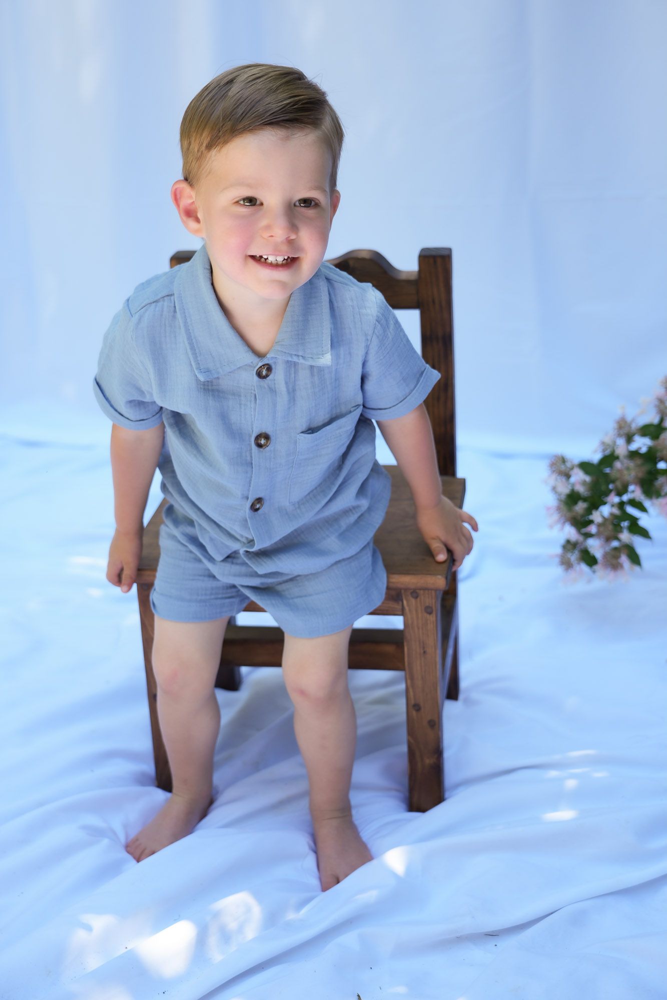 Smiling toddler boy in blue linen shorts set standing beside small wooden chair.