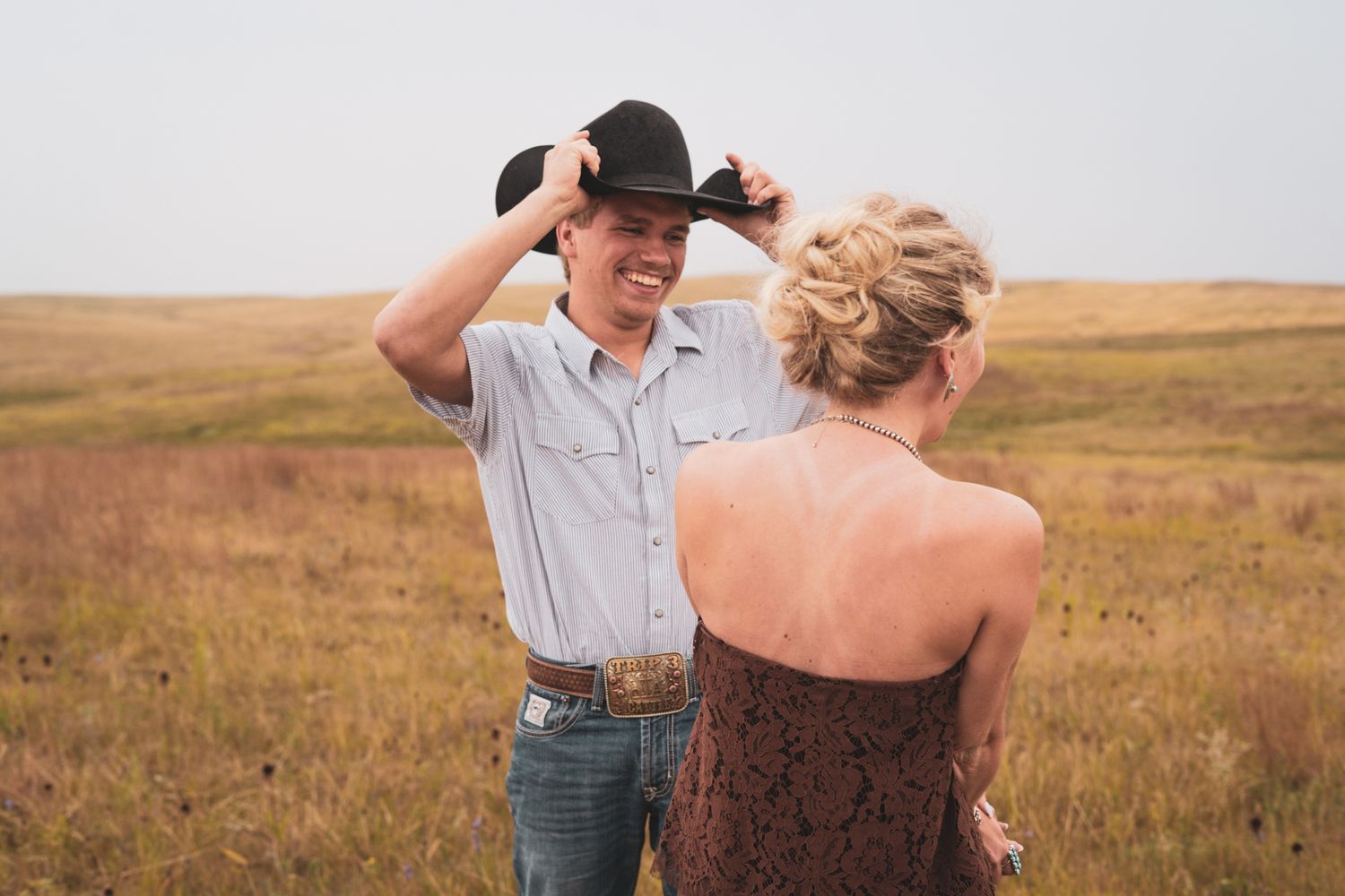 Smiling man in black cowboy hat and woman sharing a joyful moment in a wide open prairie field.