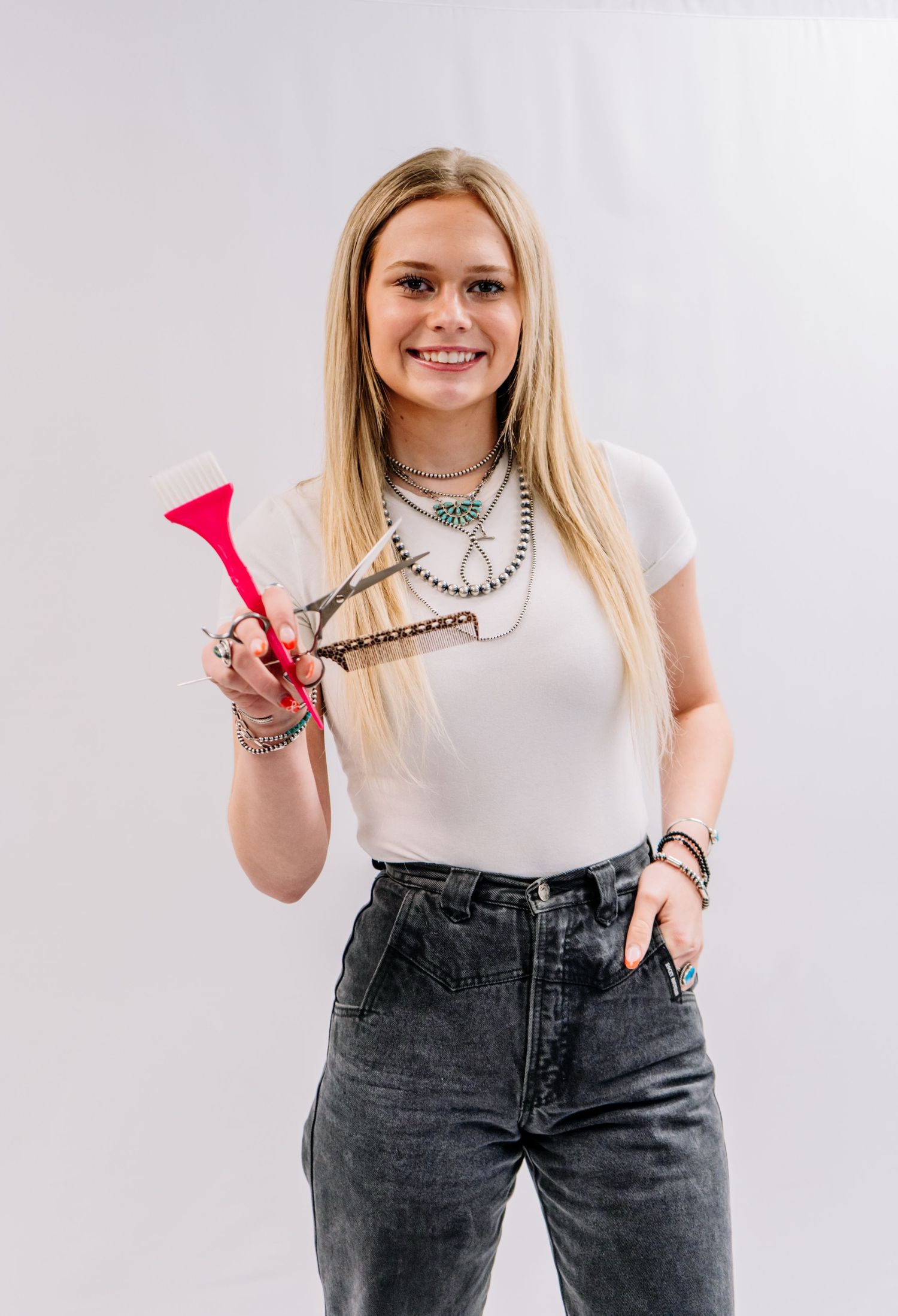 Smiling blonde woman holding red hair shears wearing white tee, gray jeans, and layered silver necklaces.