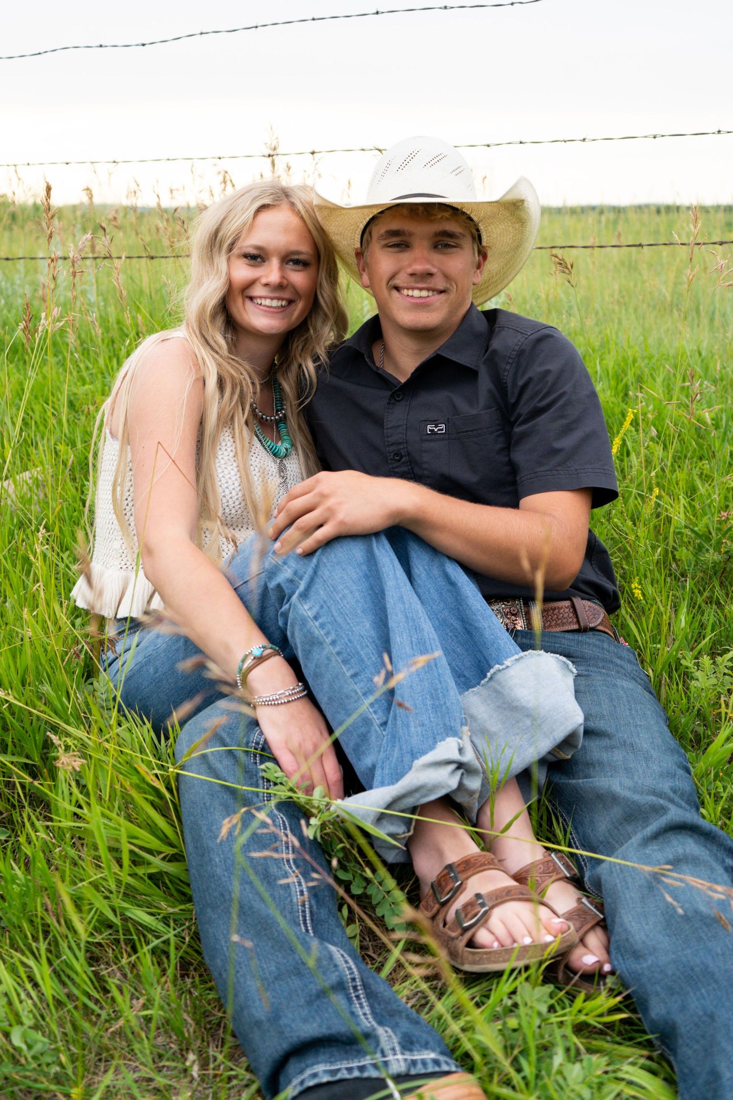 A smiling young couple sits together in tall grass near a barbed wire fence.
