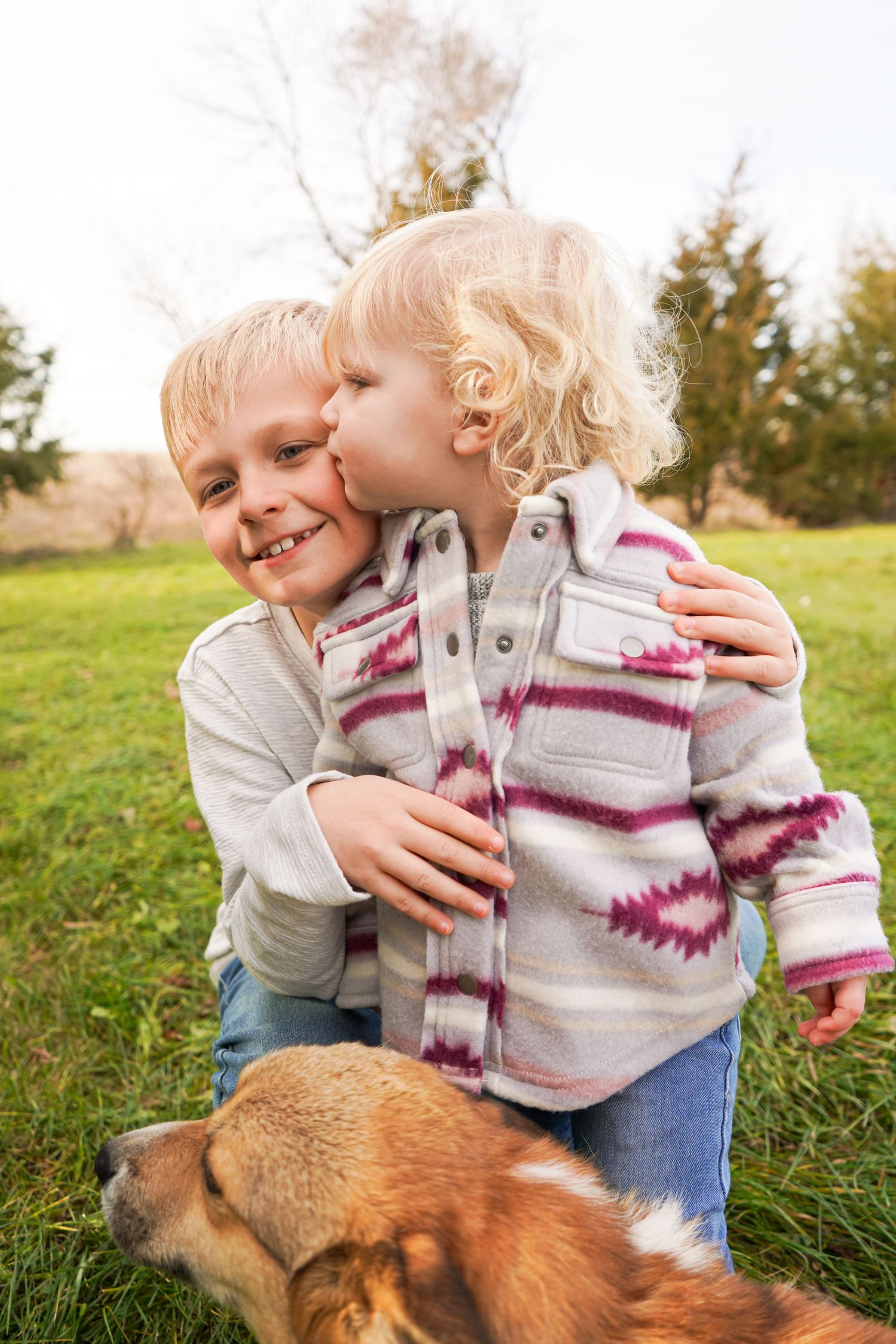 Two young blonde siblings share a sweet kiss on the cheek outdoors with a dog nearby.