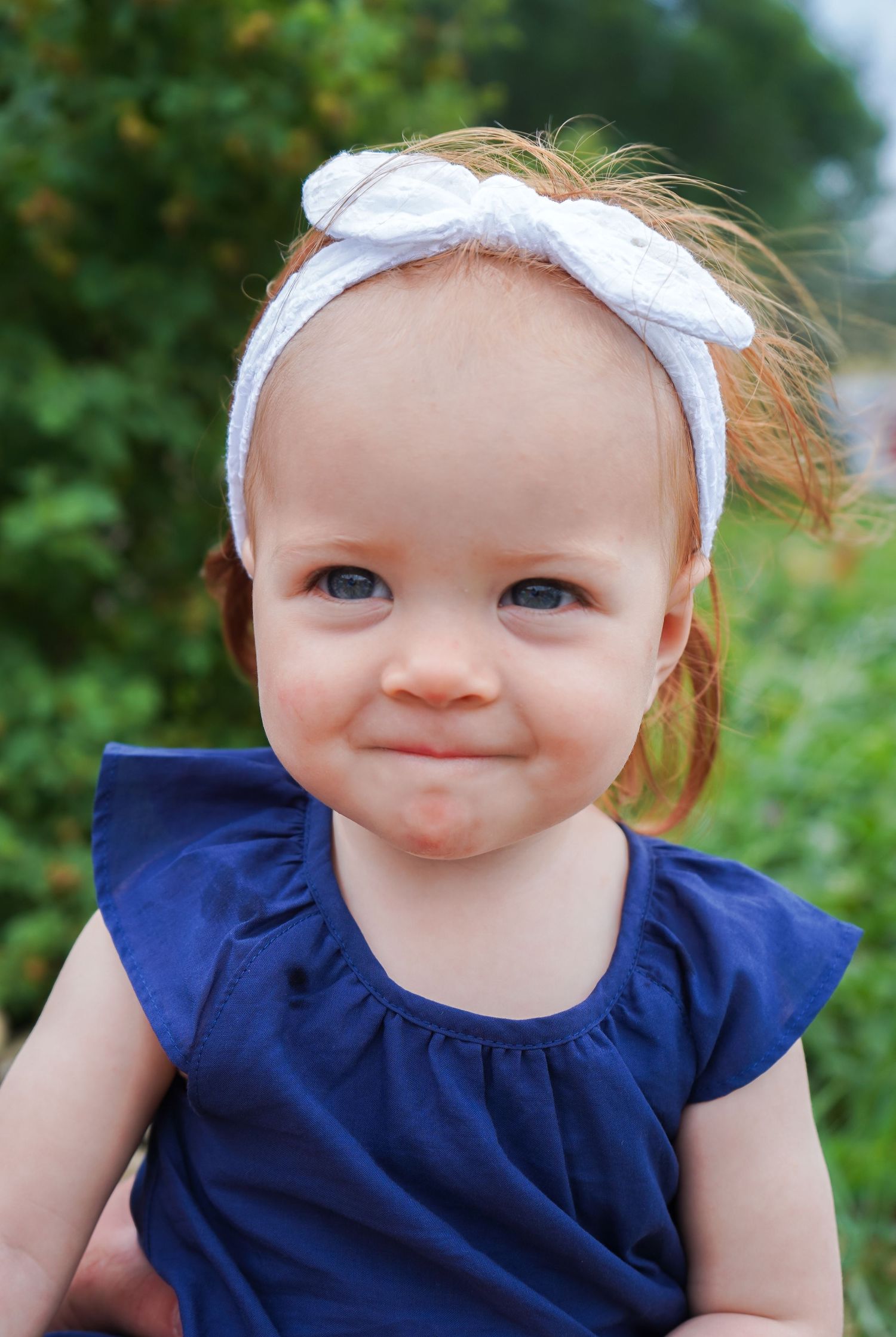 Adorable red-haired baby girl in a navy dress and white bow headband making a funny face outdoors.