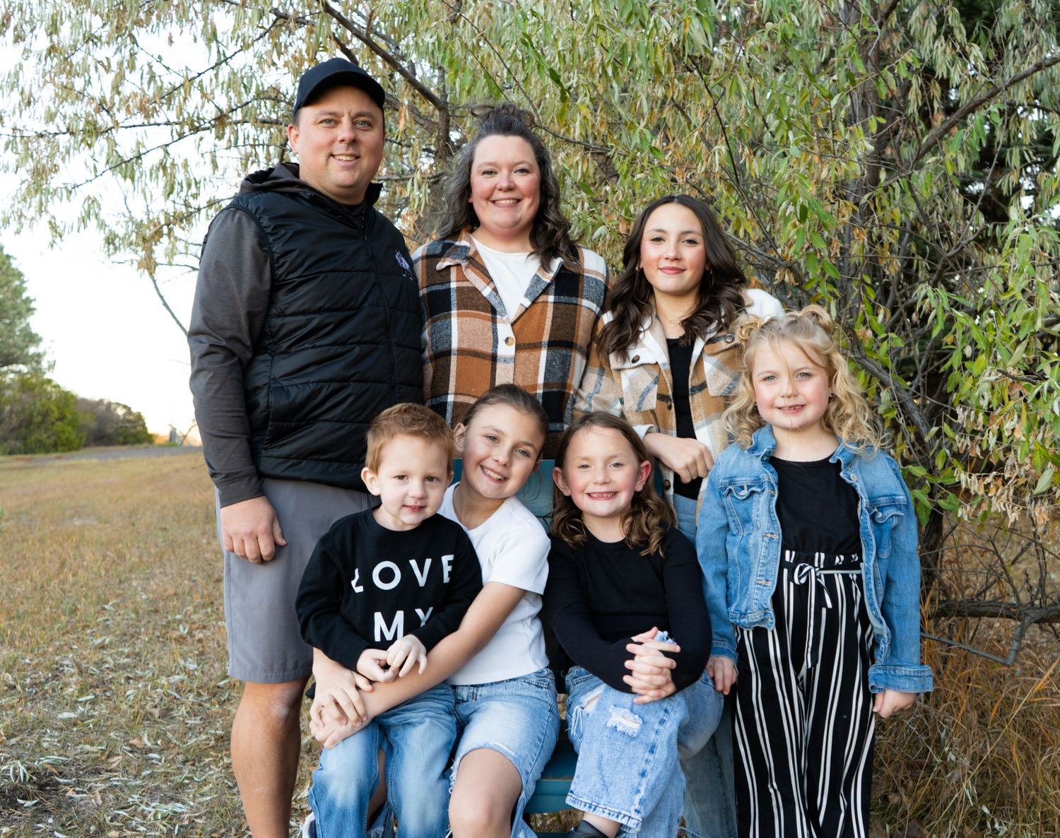Family of seven smiling together outdoors beneath a willow tree in a grassy field.