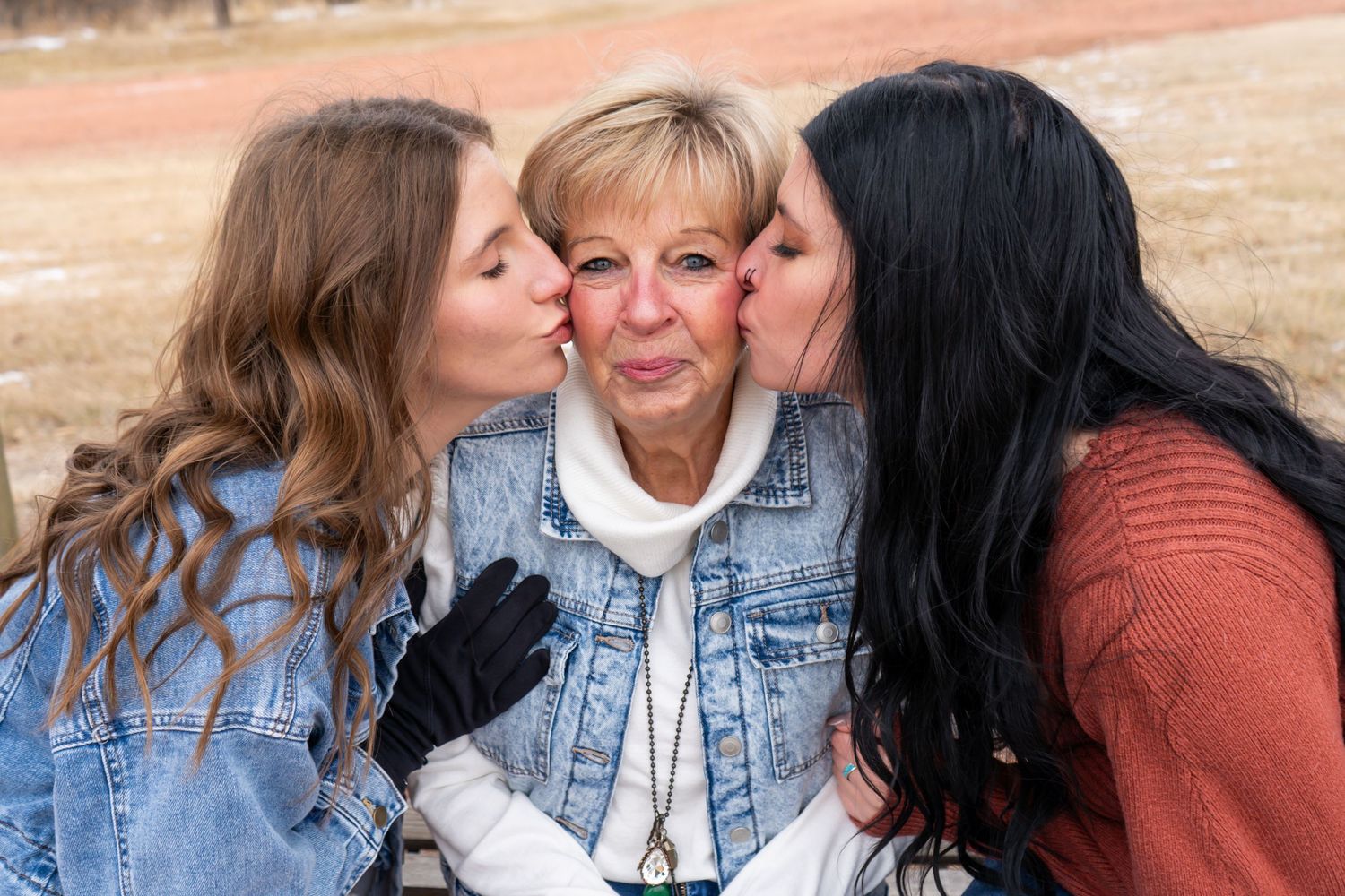 Older woman receiving kisses on cheeks from two younger women outdoors in winter setting.