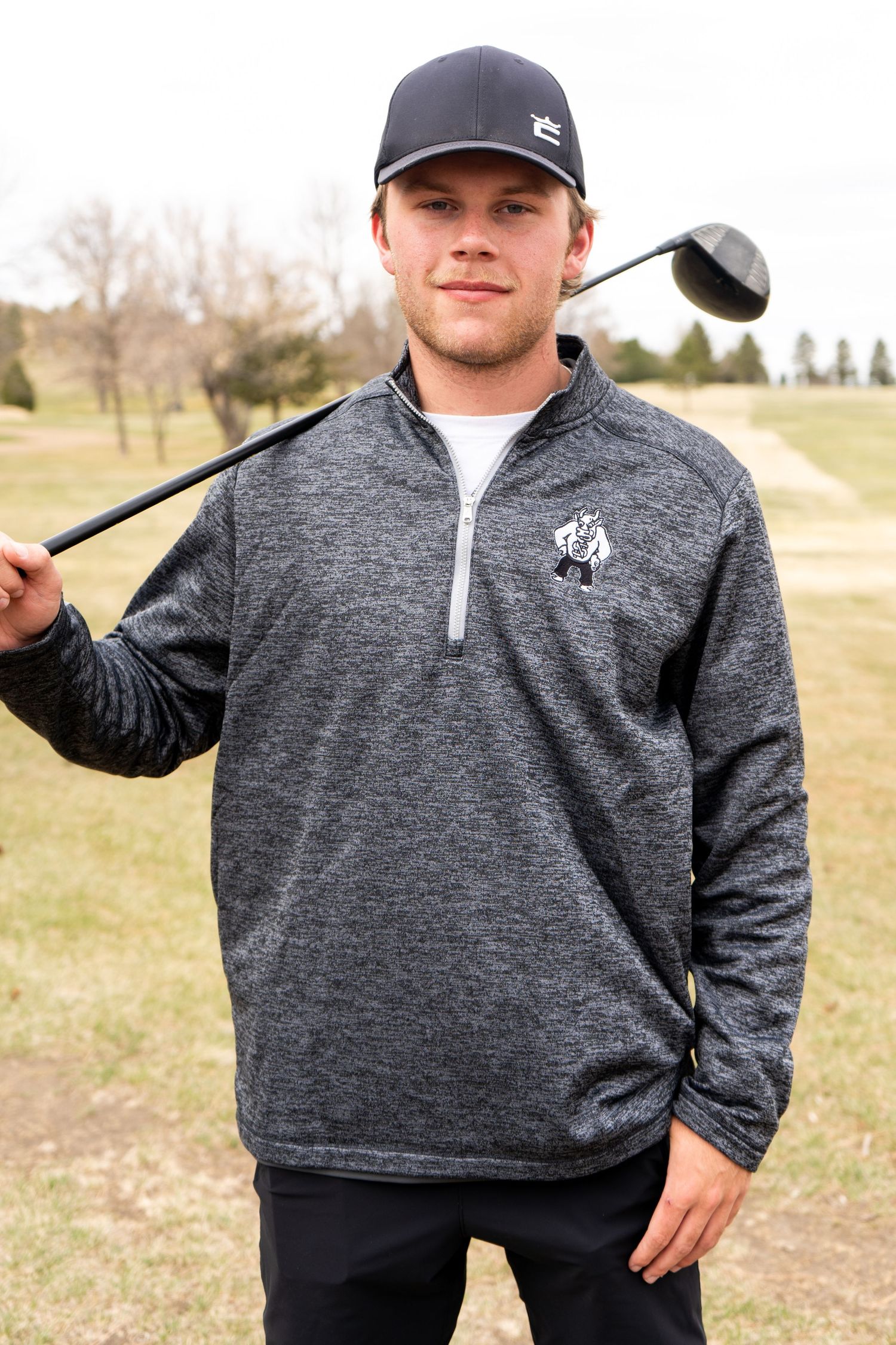 A young man in a gray quarter-zip pullover holds a golf club over his shoulder on a golf course.