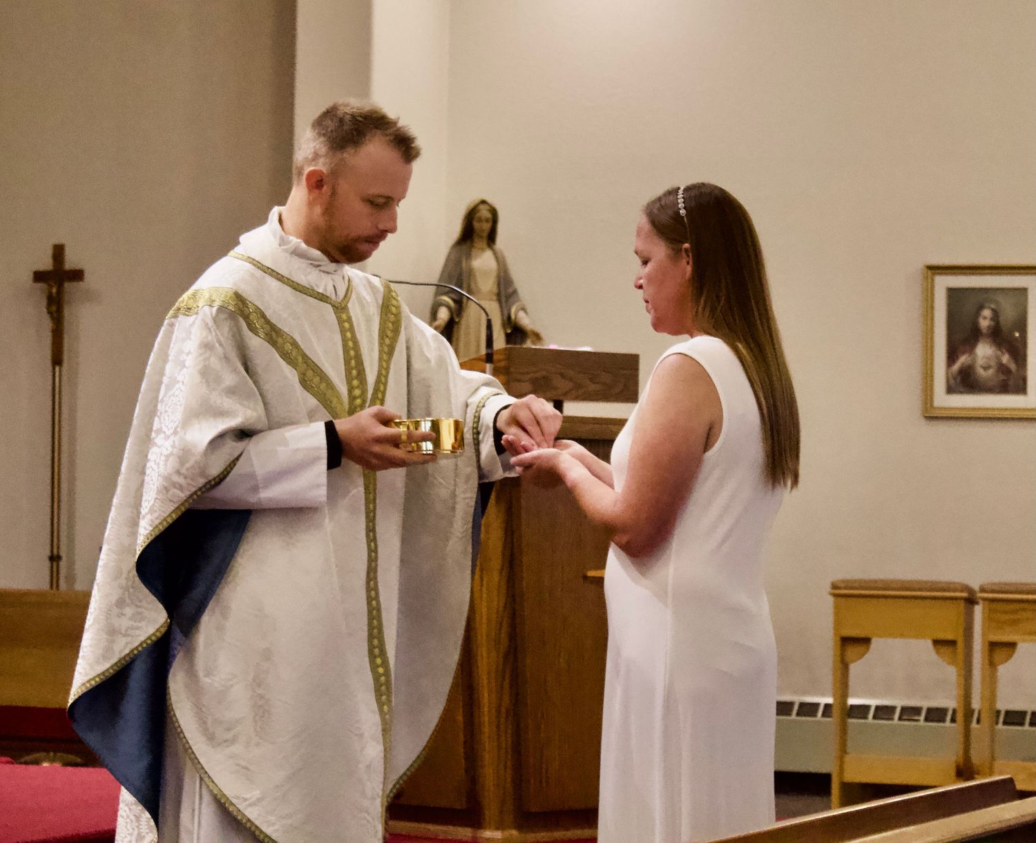 Priest offering communion to a woman in white dress during a Catholic church ceremony.