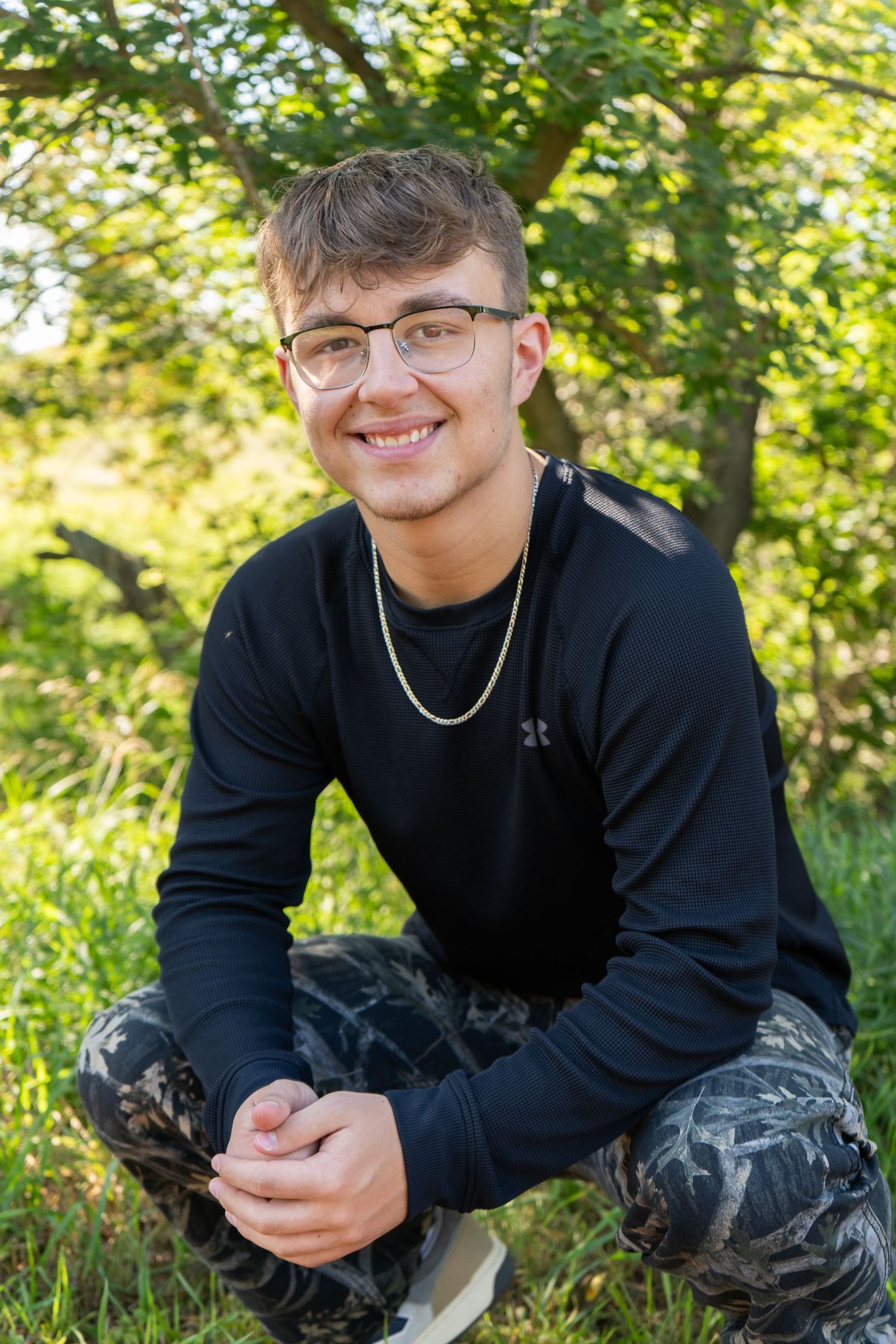 Smiling young man wearing glasses and black shirt crouching in lush green outdoor setting.