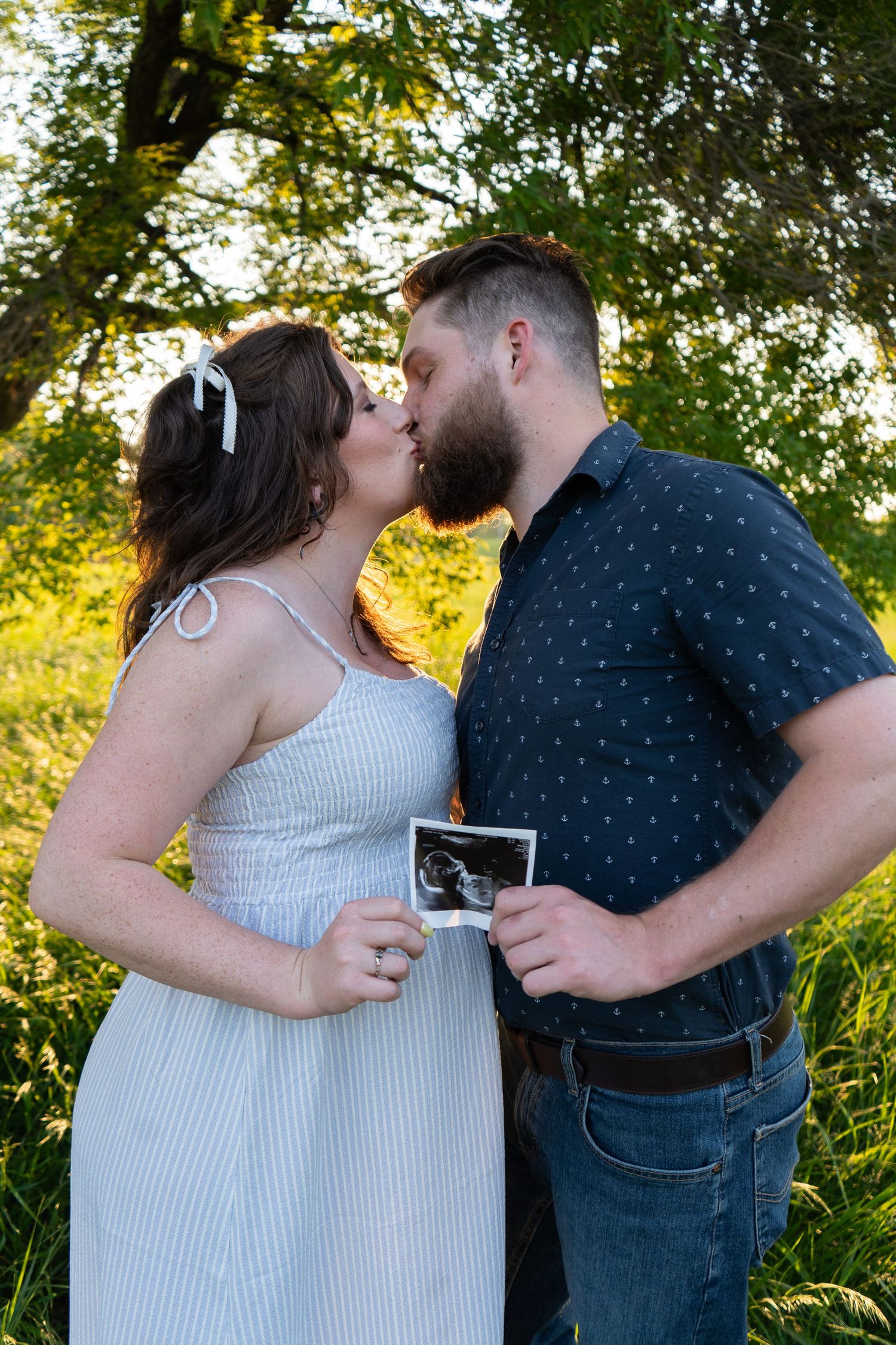 Couple kissing outdoors while holding ultrasound photo in golden sunlit garden setting.