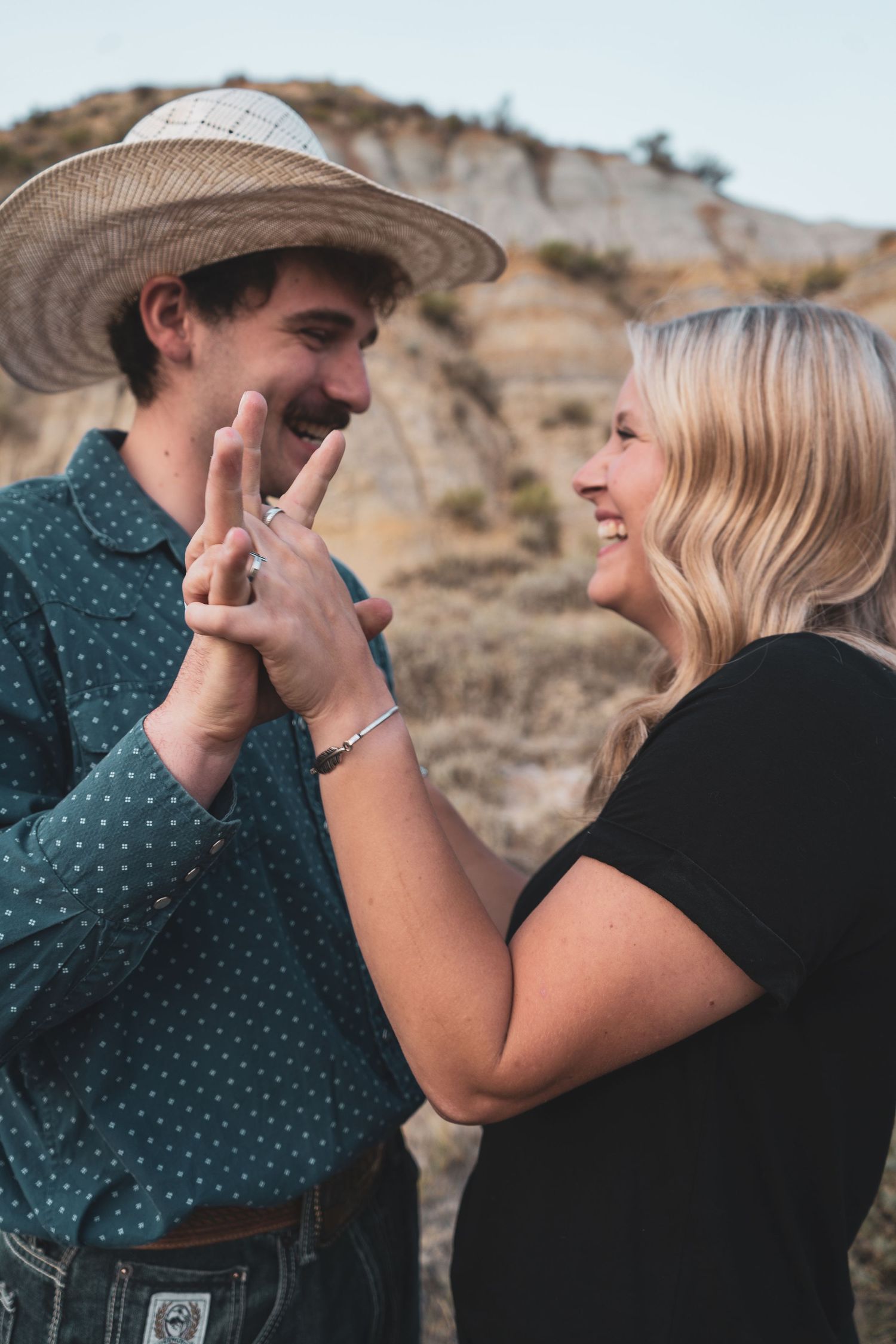 An engaged couple smiles while showing off a ring in front of rocky badlands scenery.