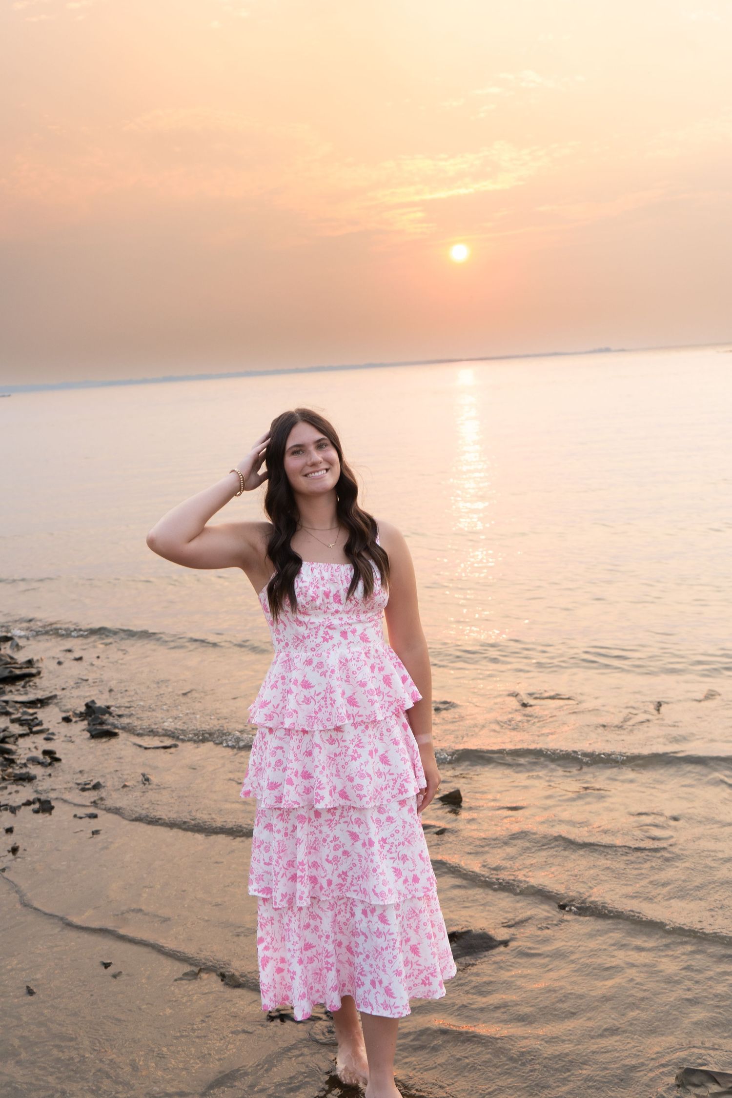 Young woman in pink floral tiered dress poses on rocky shoreline during vibrant orange sunset.