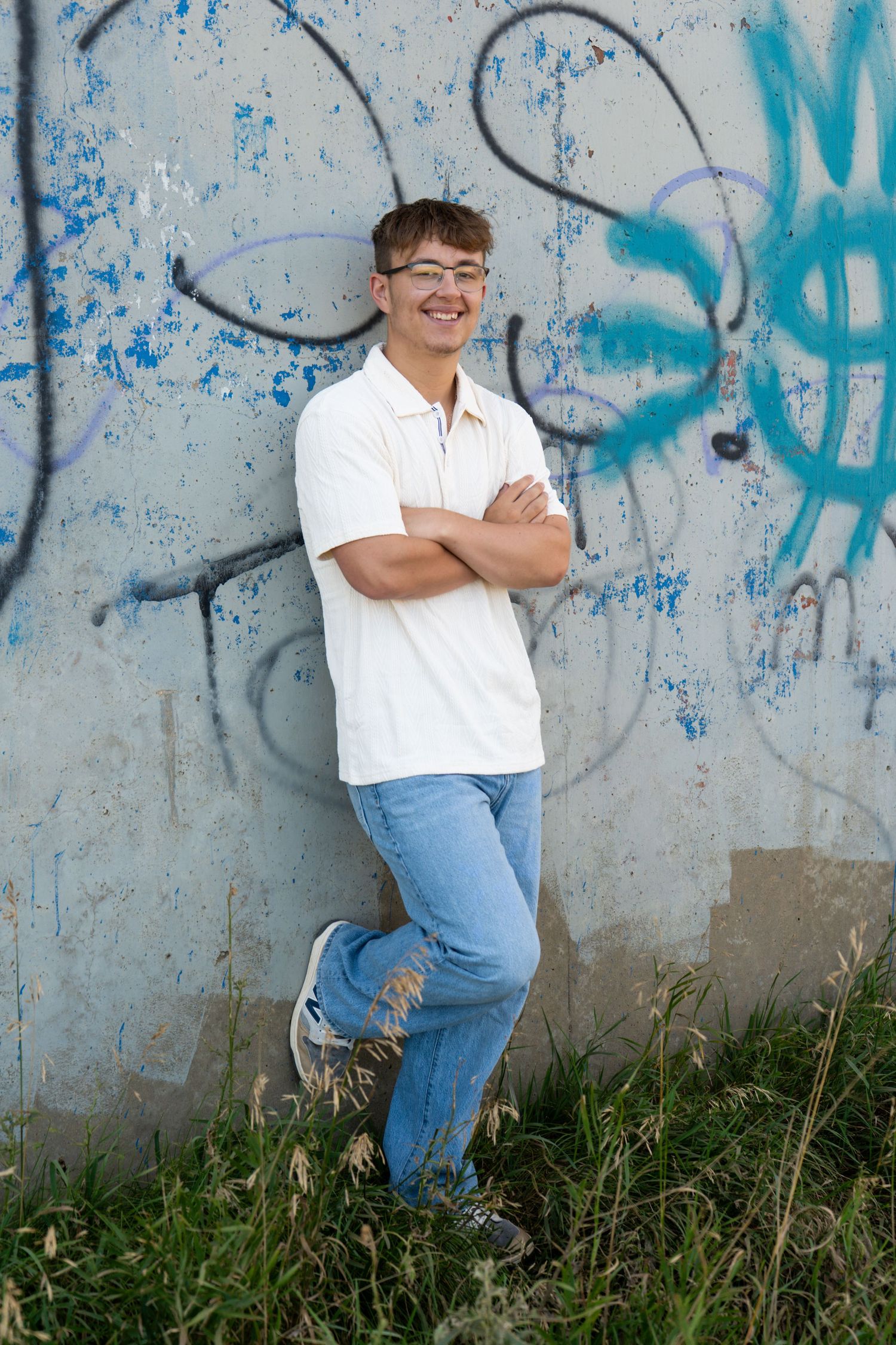 Smiling teen boy in white polo and jeans leaning against graffiti wall outdoors in color photo.