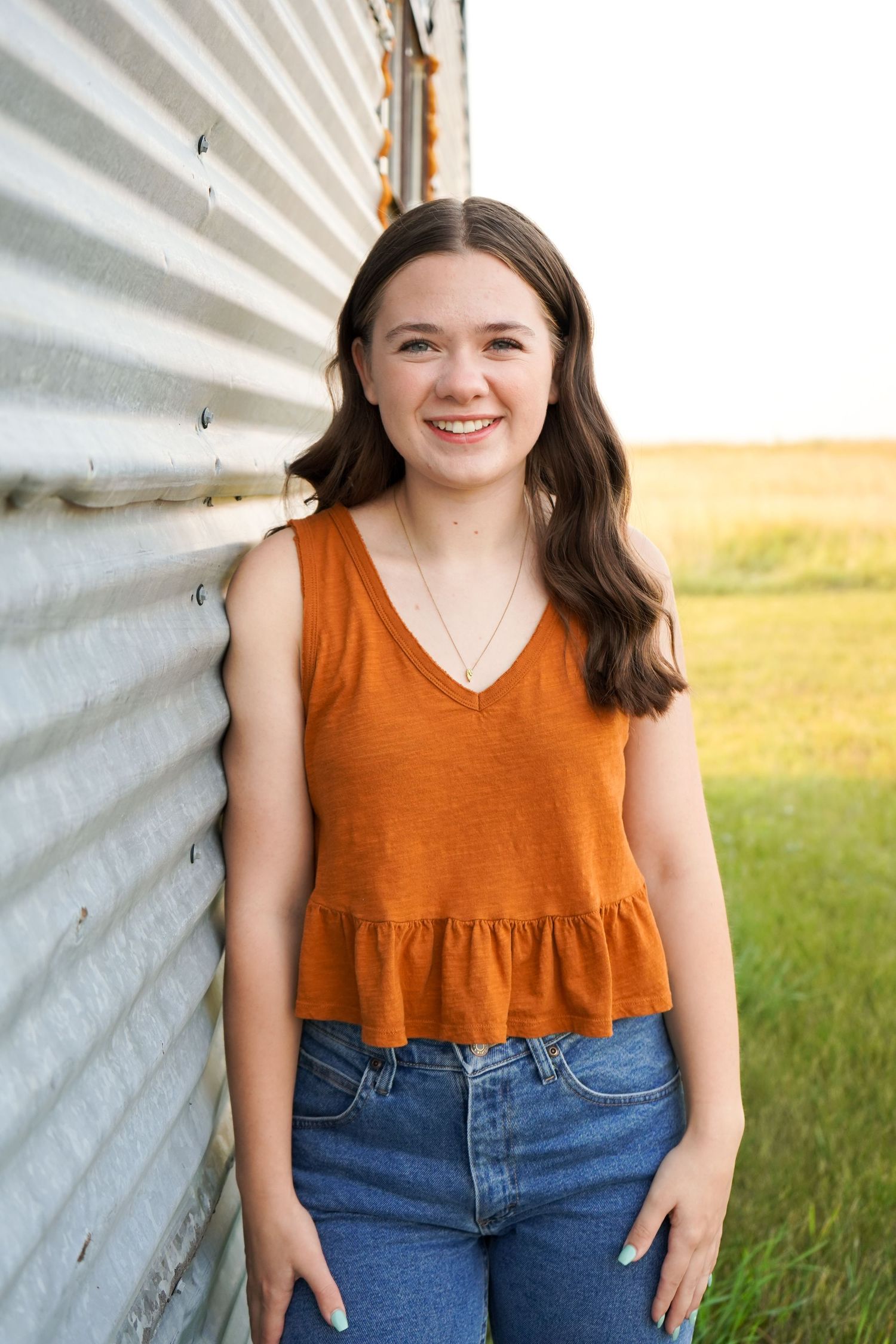 Smiling brunette teen girl in rust orange tank top leaning against corrugated metal wall.