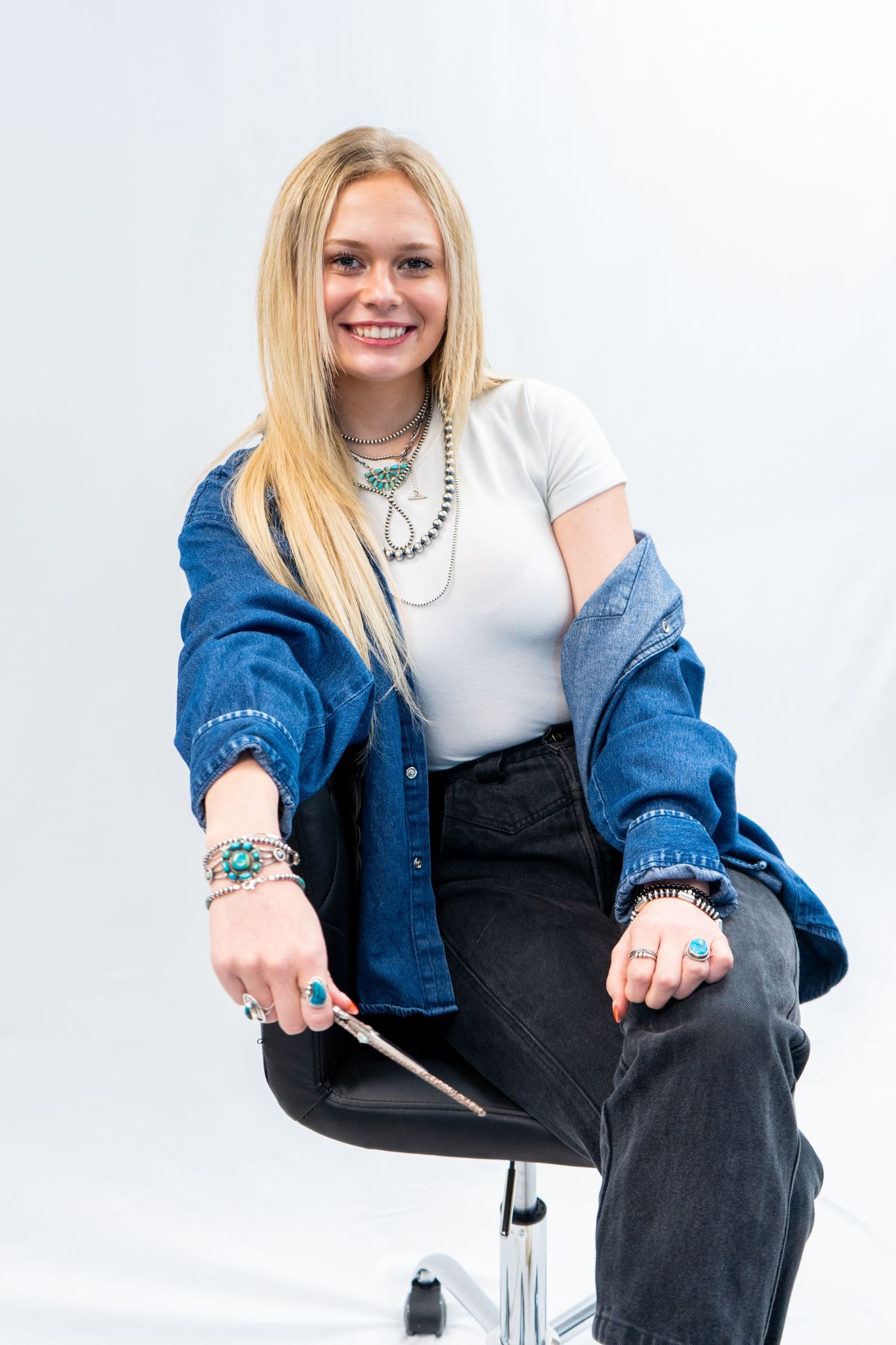 Smiling blonde woman sitting in salon chair wearing denim jacket and turquoise jewelry holding a hair comb.