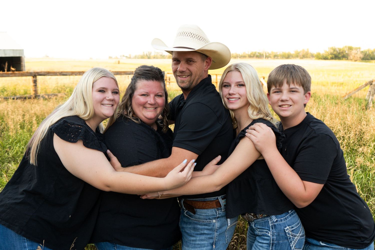 Family of five in matching black outfits embracing in a sunny farm field.