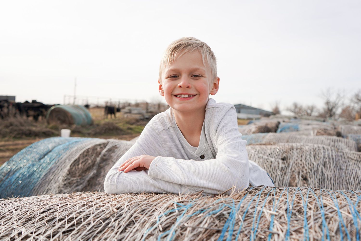 Smiling blonde boy rests on wrapped hay bales covered in blue netting on a farm.