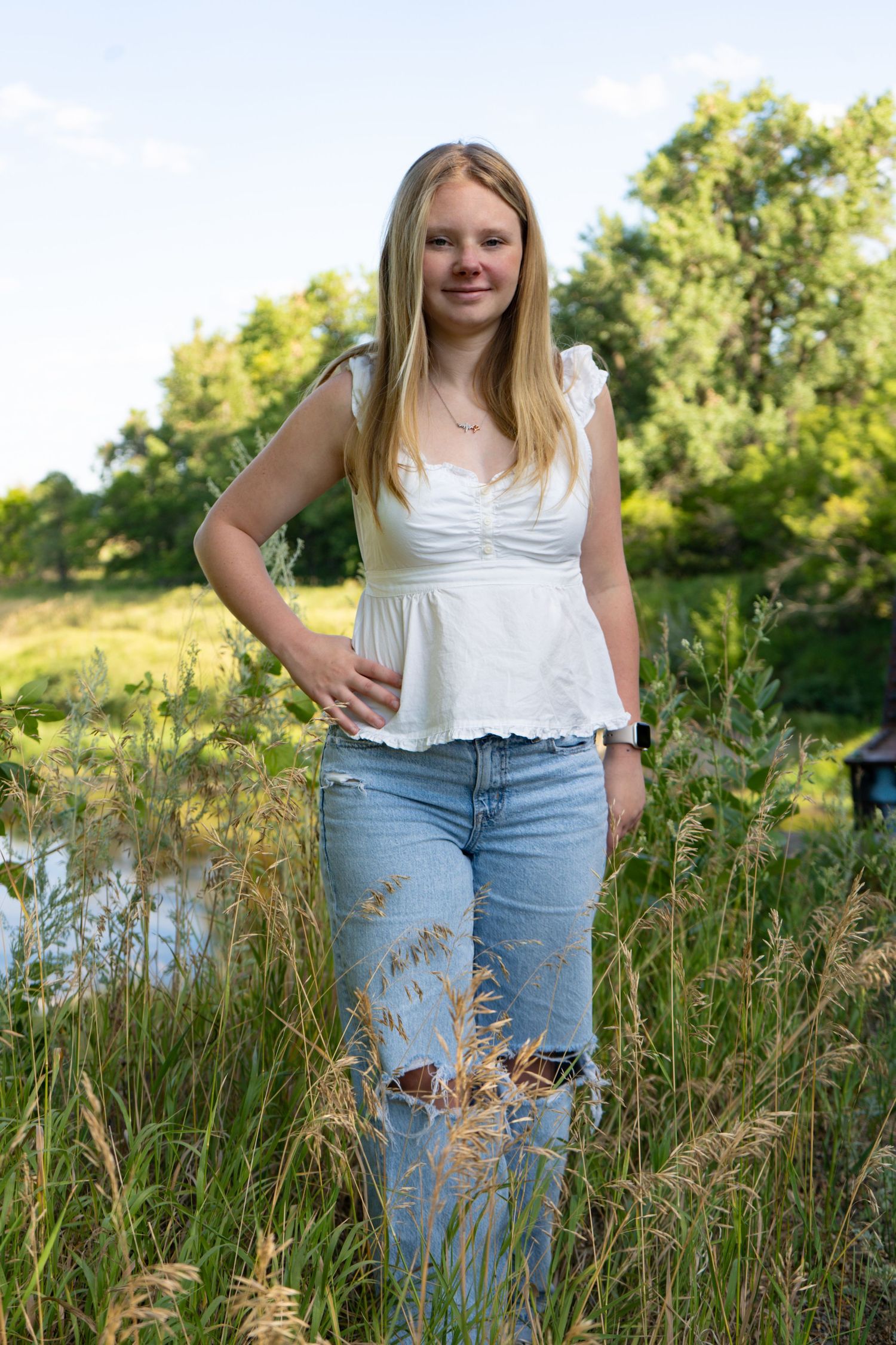 Blonde teen girl in white top and ripped jeans stands in tall grass by water.