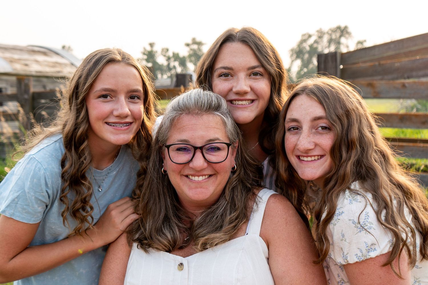A mother and her three teenage daughters smile together in a warm outdoor portrait.