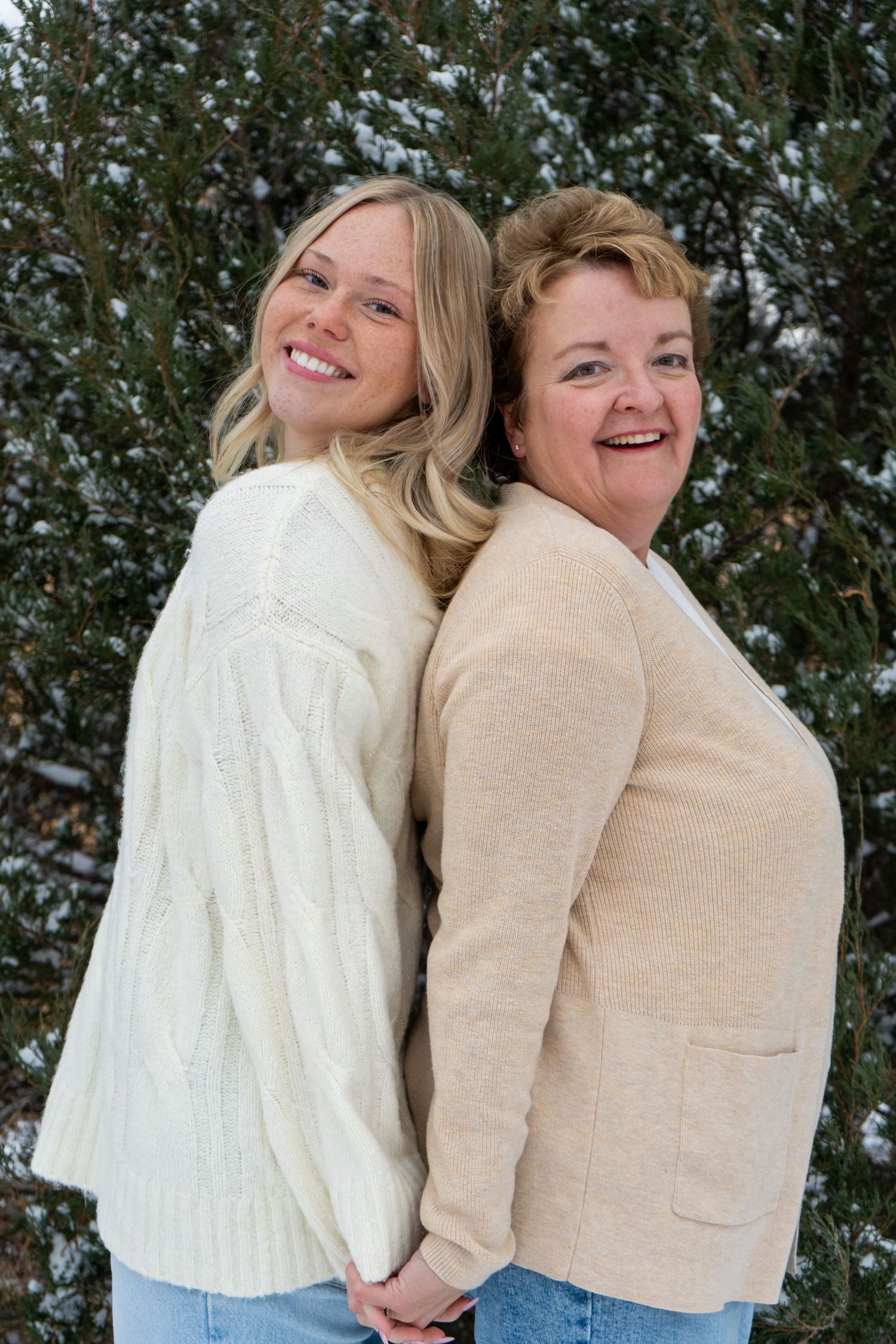 Two women standing back-to-back smiling in front of snow-dusted evergreen trees in winter.