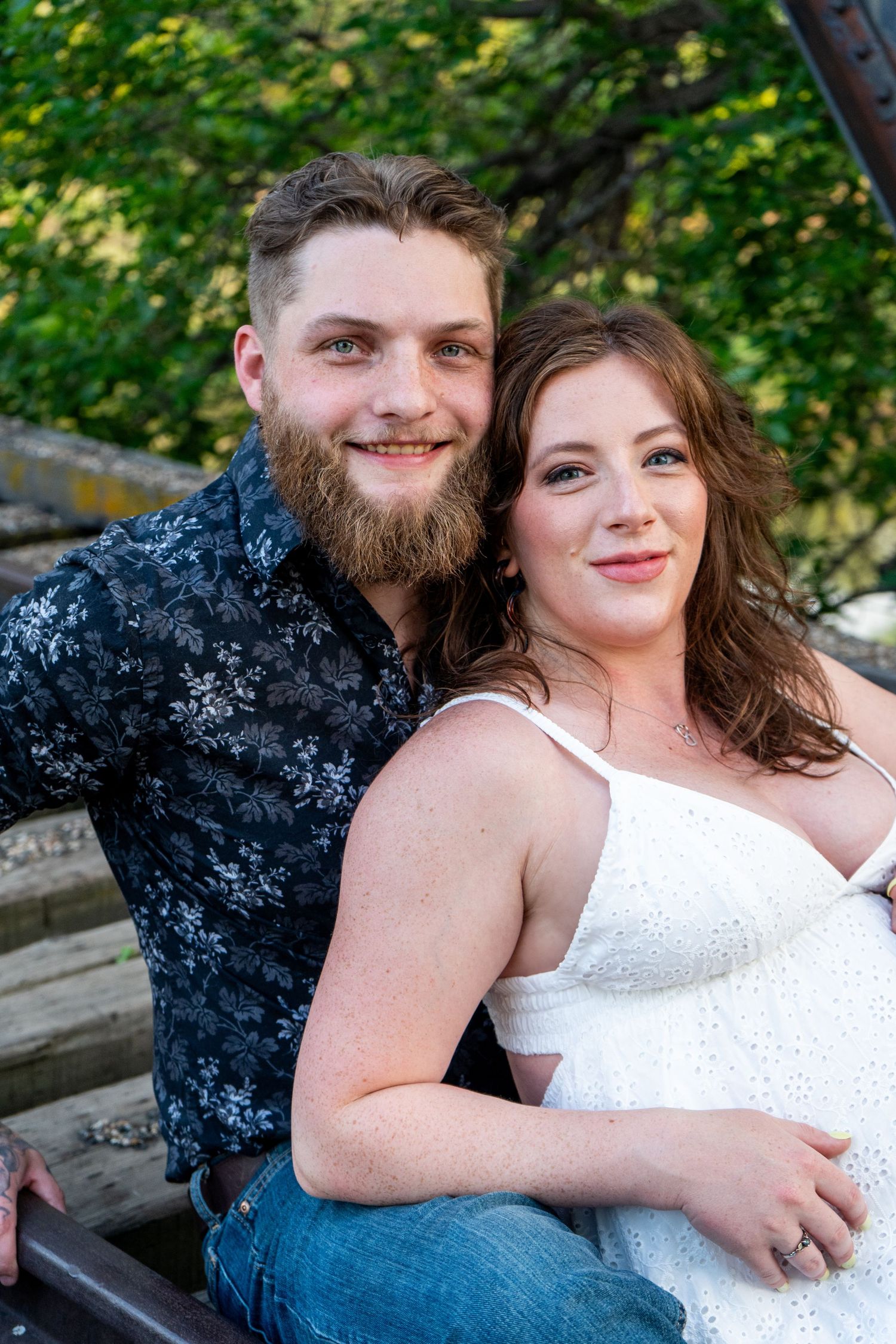 Smiling couple sitting together outdoors, man with beard in floral shirt, woman in white dress.
