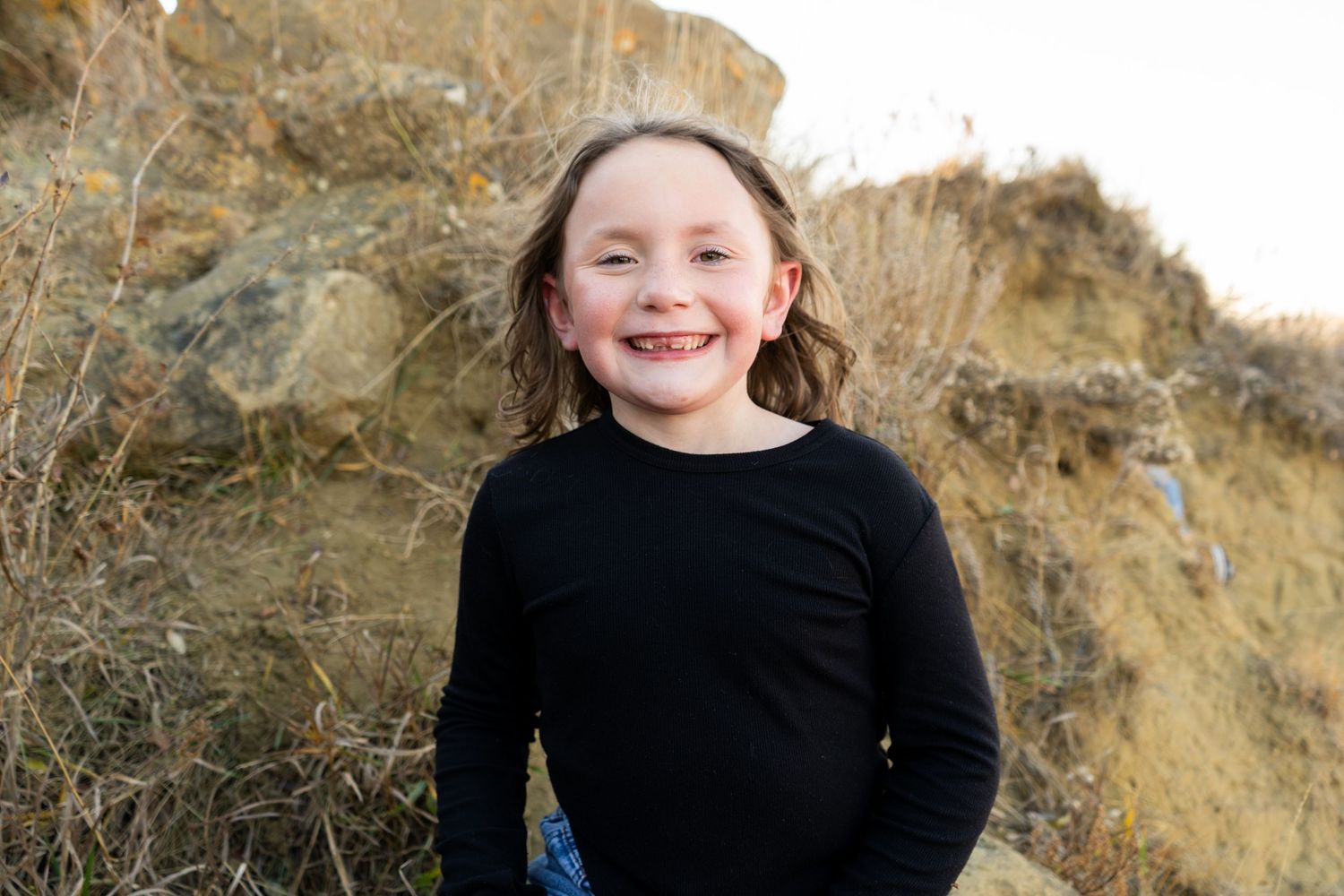 Happy young girl with curly brown hair smiling outdoors wearing black long-sleeve shirt.