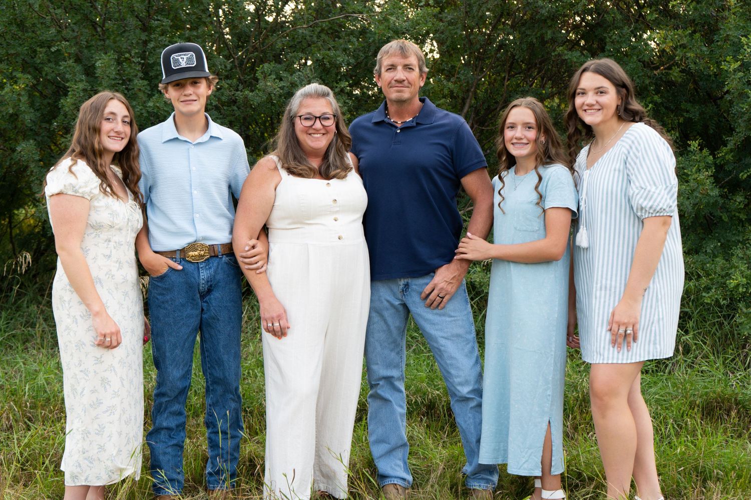 Family of six in coordinating outfits smiles together outdoors surrounded by green trees.