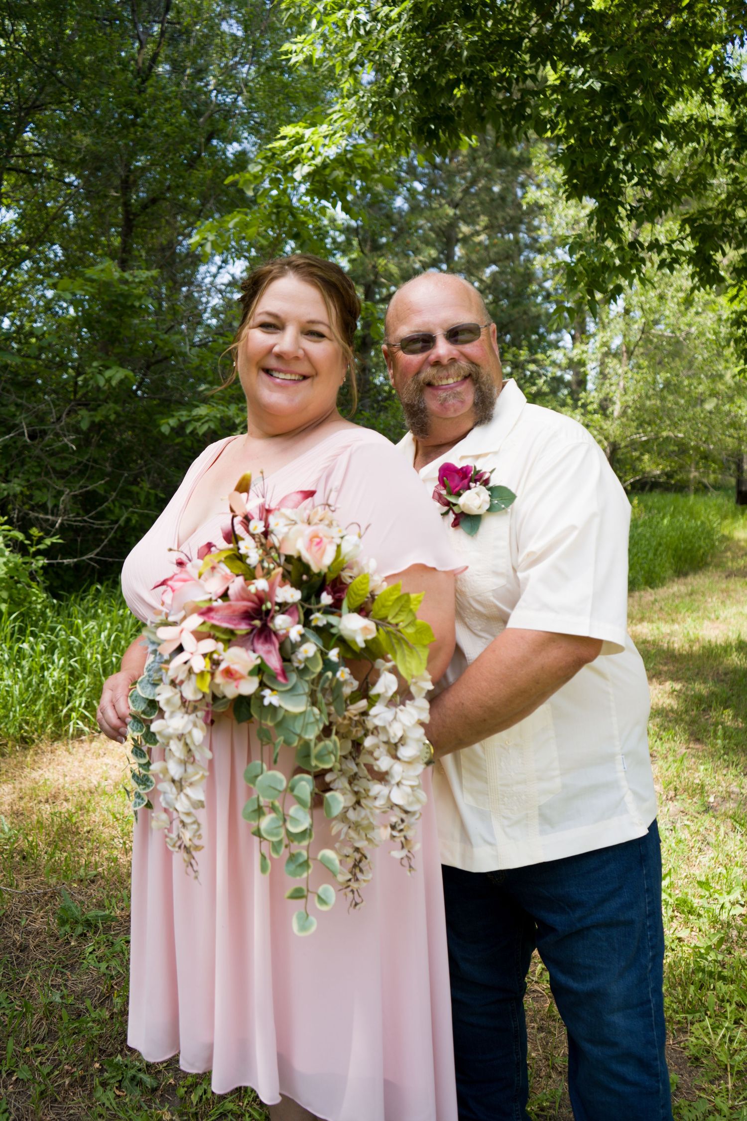 Smiling wedding couple outdoors, bride in pink gown holding cascading bouquet, groom in cream shirt with boutonniere.