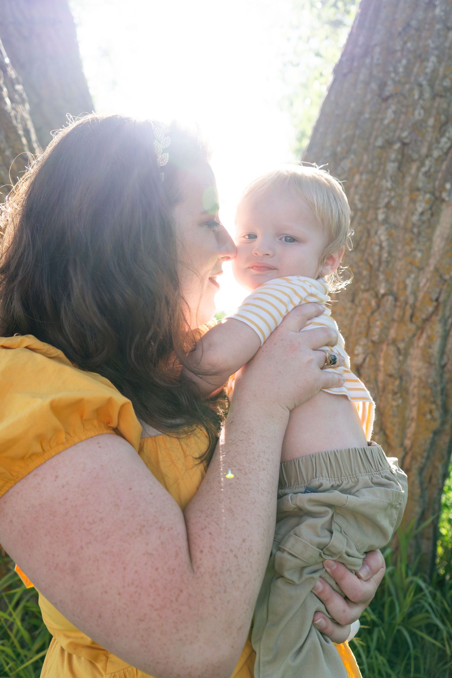 A mother tenderly kisses her baby outdoors near trees in warm sunlight.