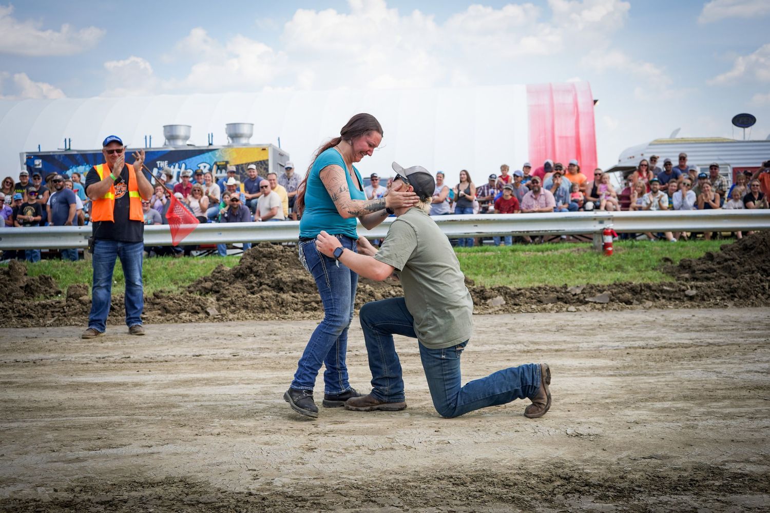 A man proposes on one knee to a woman on a dirt track in front of a large crowd of spectators.
