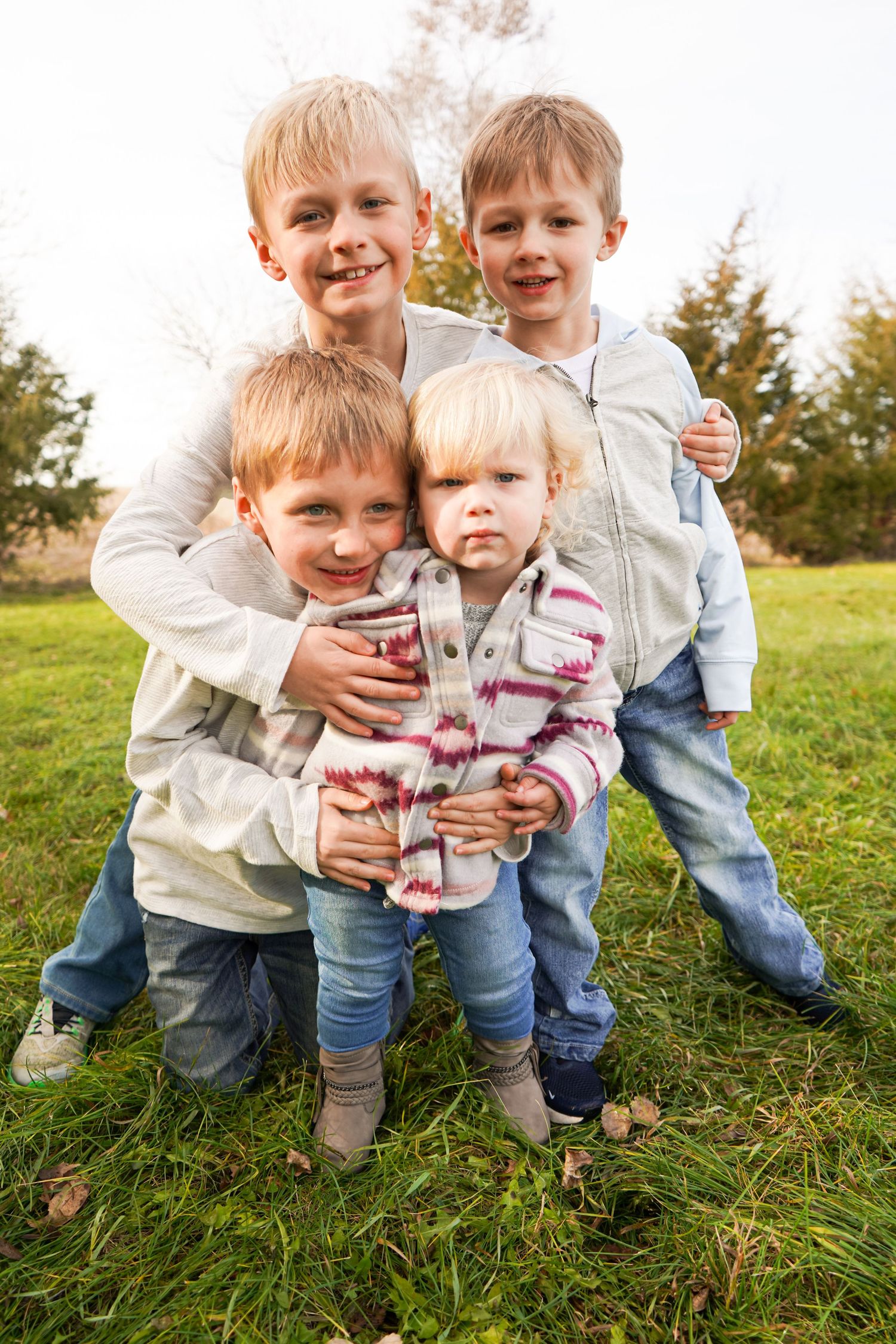 Four young siblings huddle together smiling for a group photo on a grassy lawn outdoors.