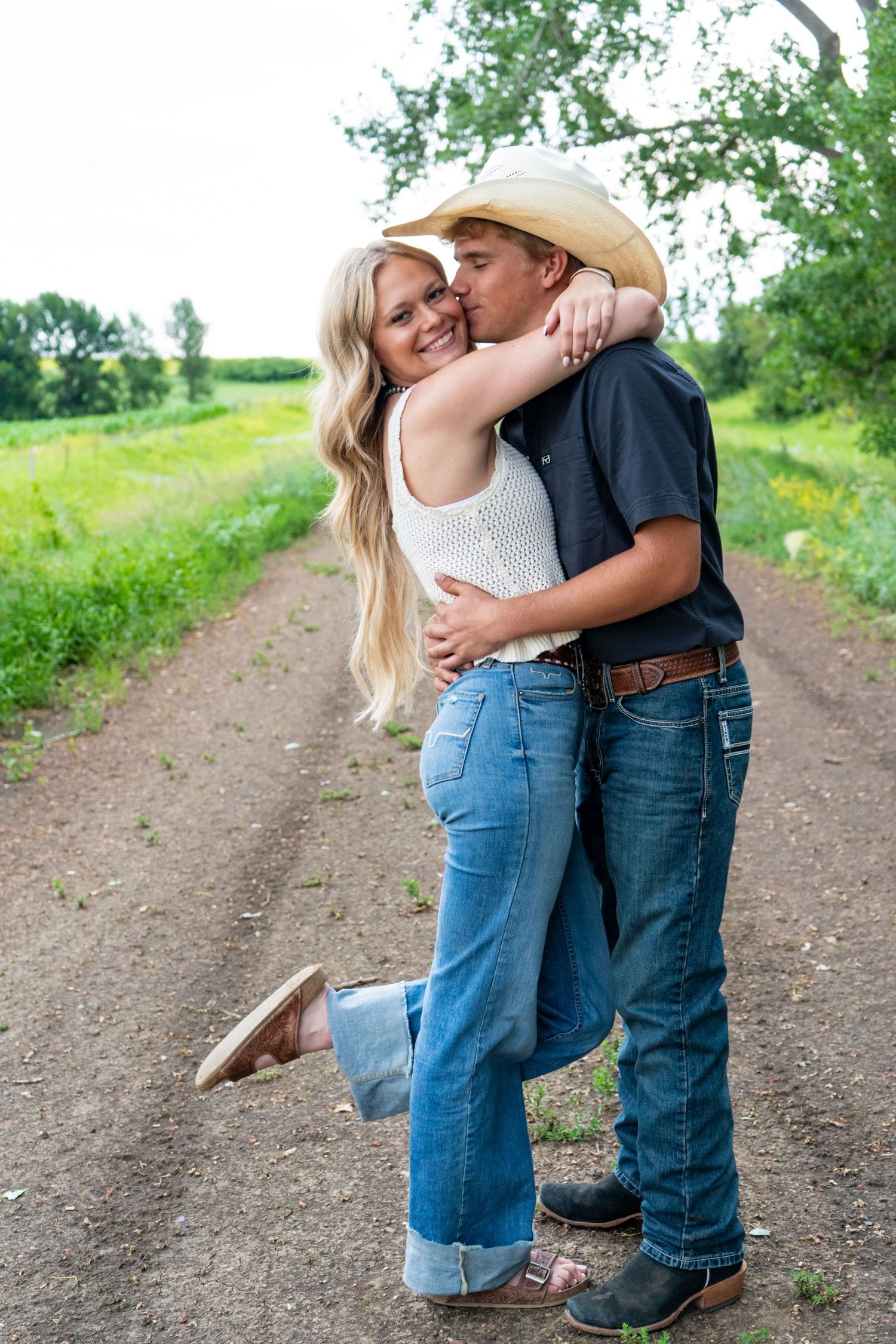 Cowboy in straw hat kissing smiling blonde woman on a scenic country dirt road.
