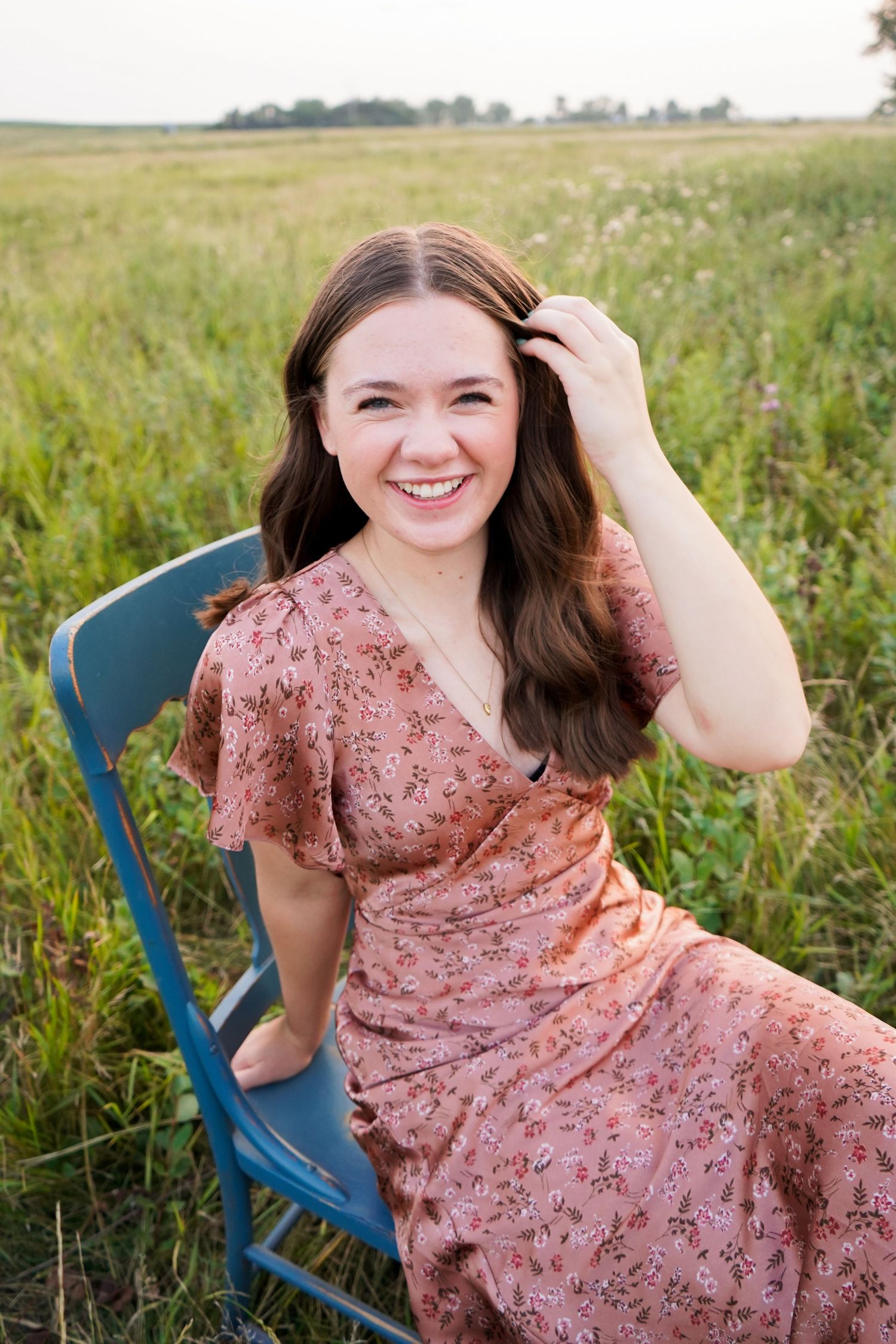 Laughing brunette teen girl in pink floral dress sitting on blue chair in green field.