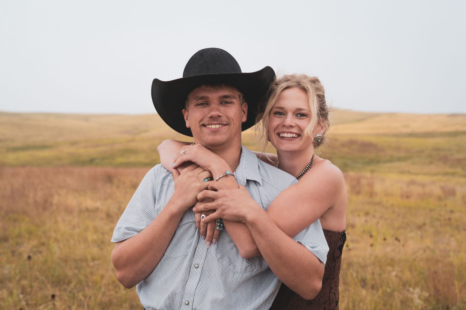A smiling blonde woman hugs a man in a black cowboy hat from behind in an open golden field.