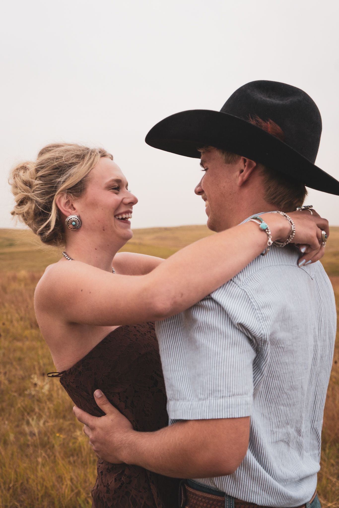 A laughing blonde woman embraces a man in a black cowboy hat in a golden field.