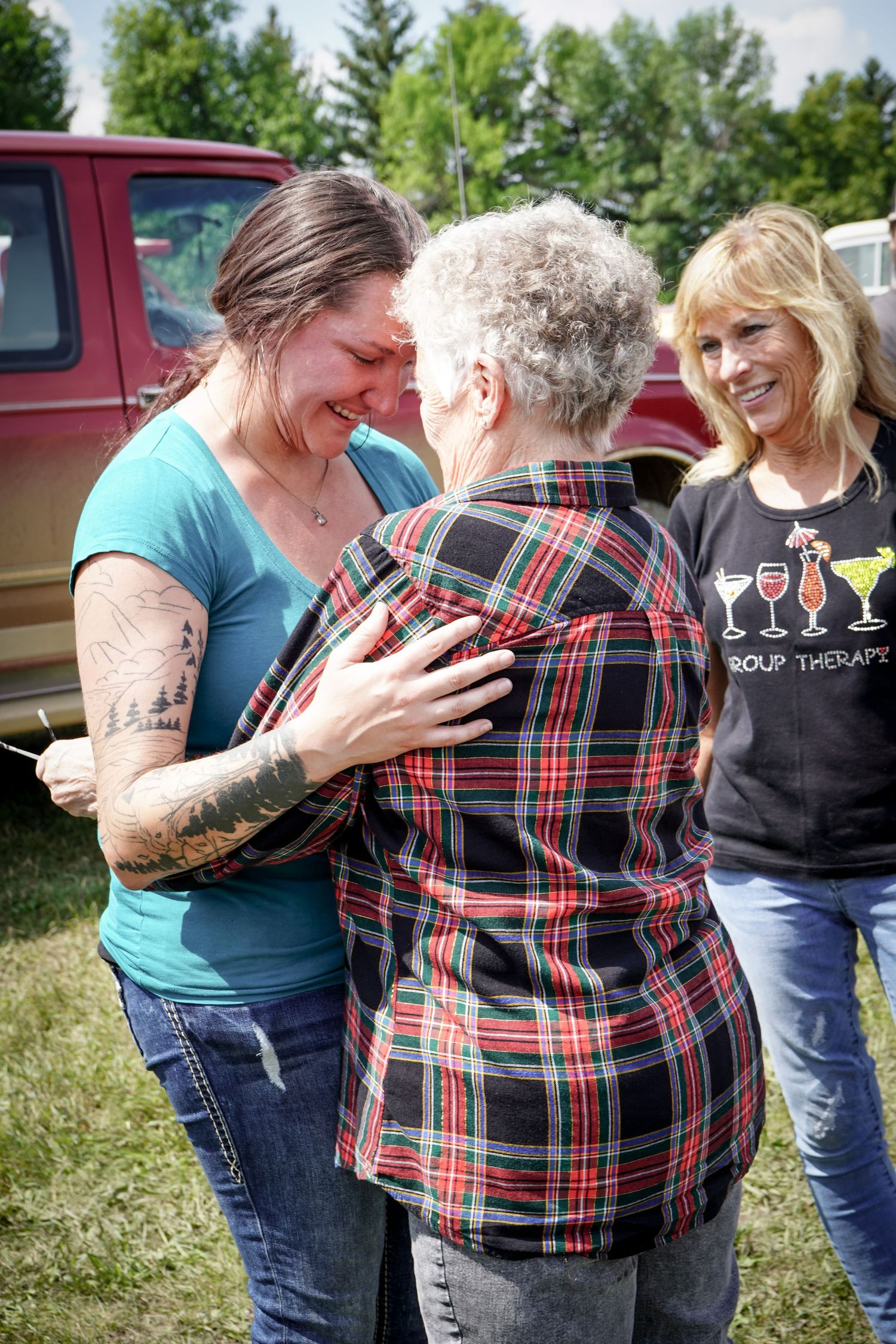 Emotional woman with tattoos hugs elderly lady while smiling woman watches outdoors.