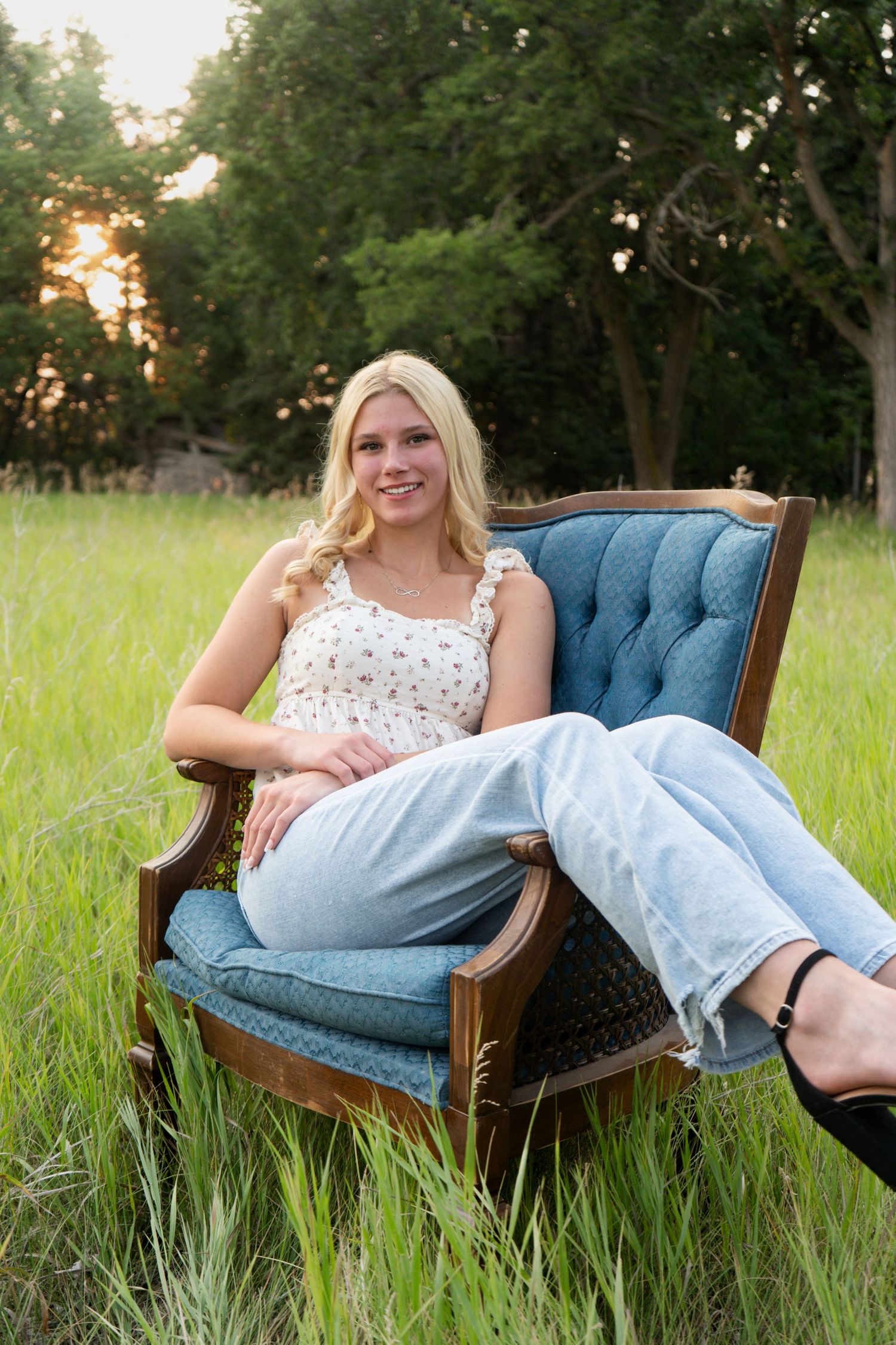 Blonde teen girl relaxing in vintage blue velvet chair in lush green grass field.