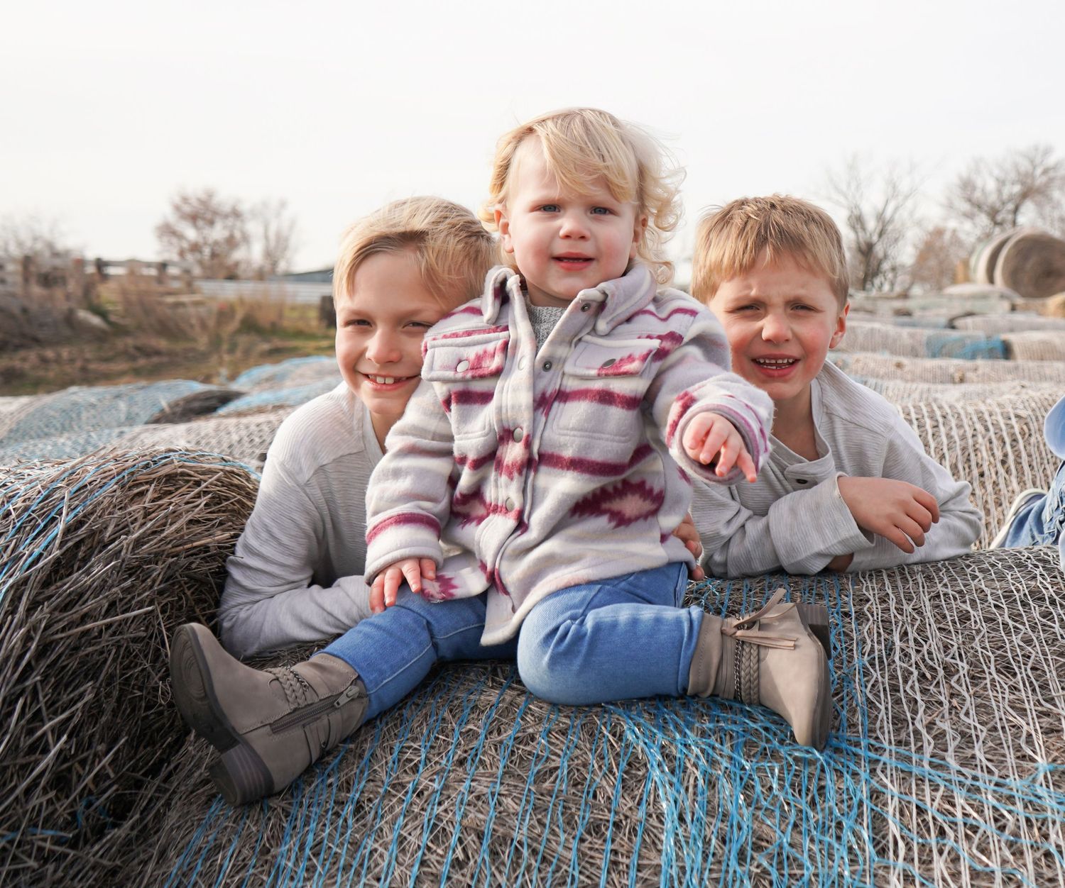 Three young siblings sit together laughing on hay bales wrapped in blue netting outdoors.