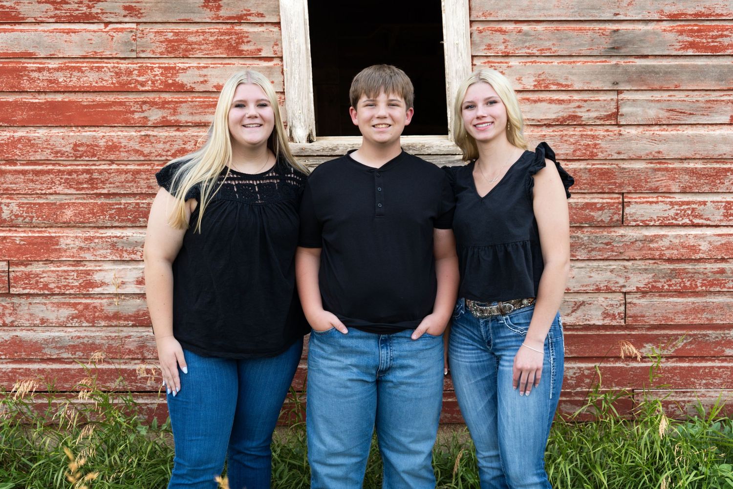 Three siblings in black tops and jeans pose smiling in front of a rustic red barn.