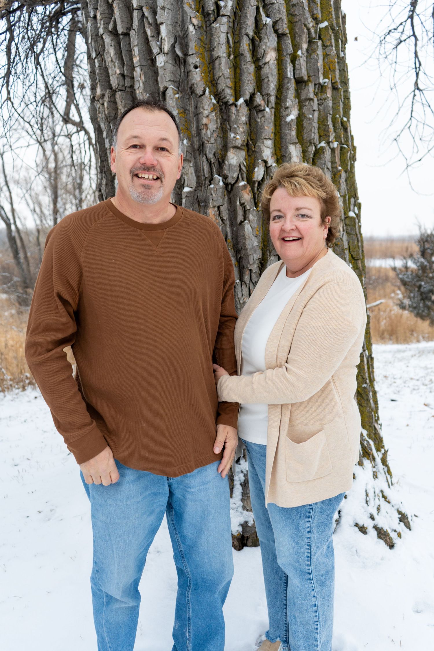 Middle-aged couple smiling outdoors in winter snow beside large tree, woman in beige cardigan, man in brown sweater.