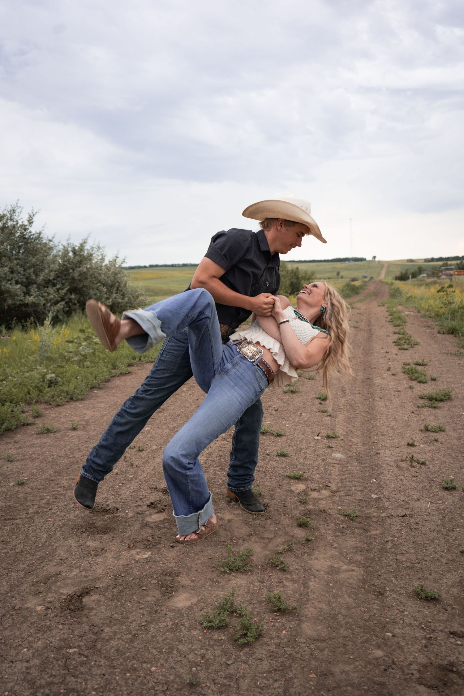 Playful couple dancing on a rural dirt road, man in cowboy hat dipping smiling blonde woman.